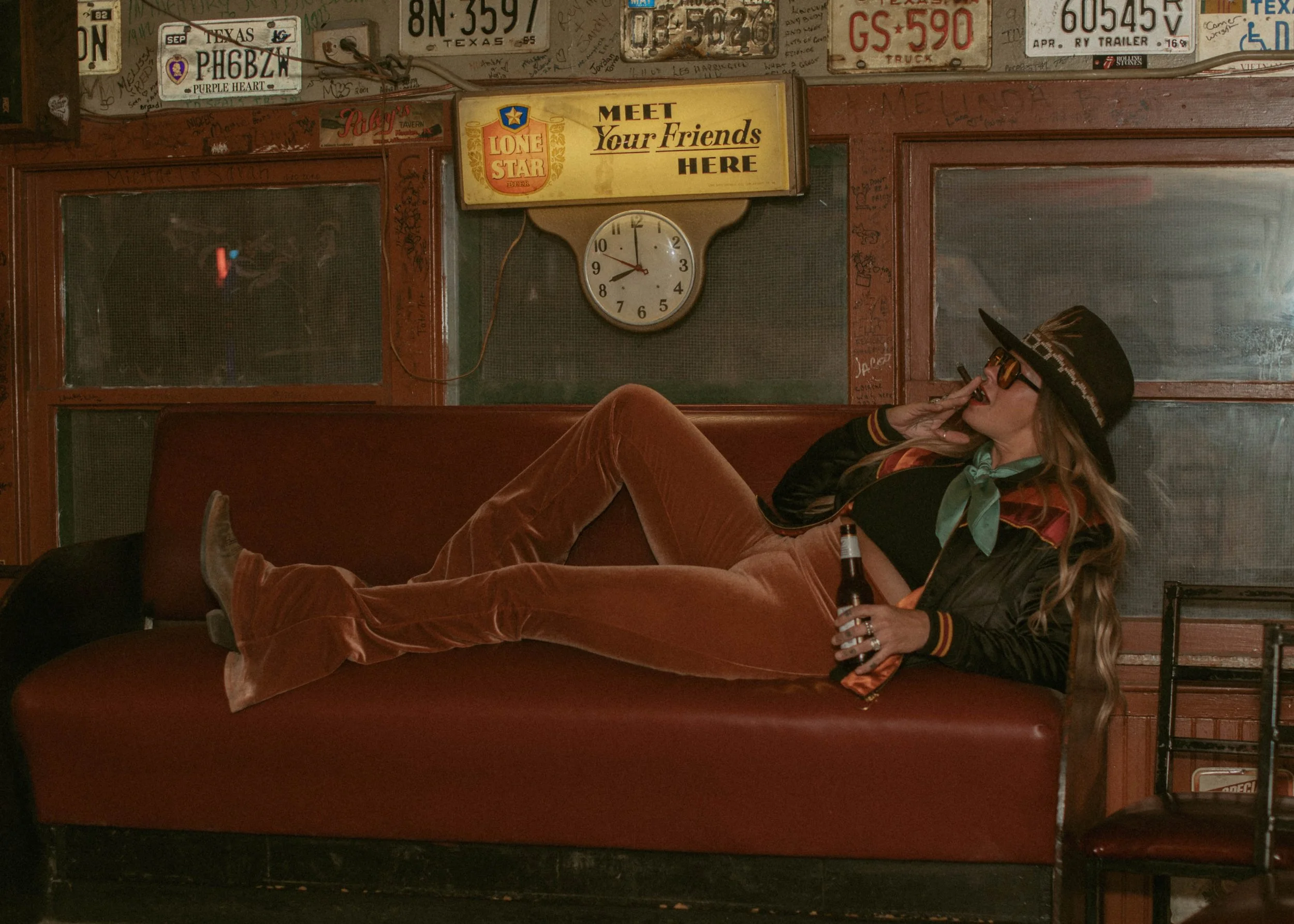 A woman sitting on a red velvet couch in a bar. She is wearing sunglasses, a black hat, and a colorful scarf. She is holding a beer bottle and has her legs crossed. The background has various license plates and signs, including a yellow sign that says "Meet Your Friends Here" and a clock showing approximately 9:50.