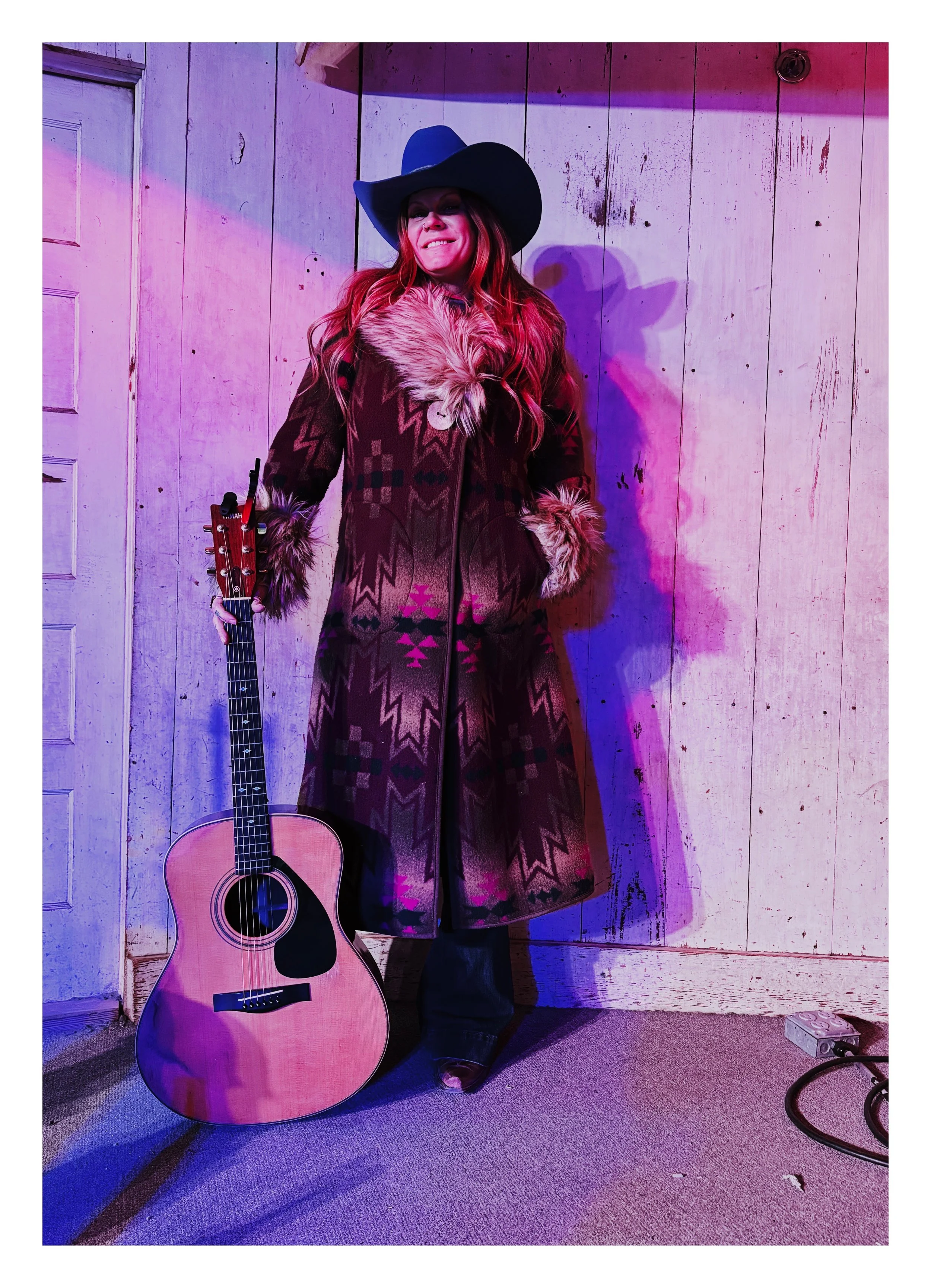 Woman with long red hair wearing a black cowboy hat and a southwestern patterned coat, standing indoors with purple and pink lighting, holding an acoustic guitar, against a wooden wall with an outlet and a power strip on the floor.