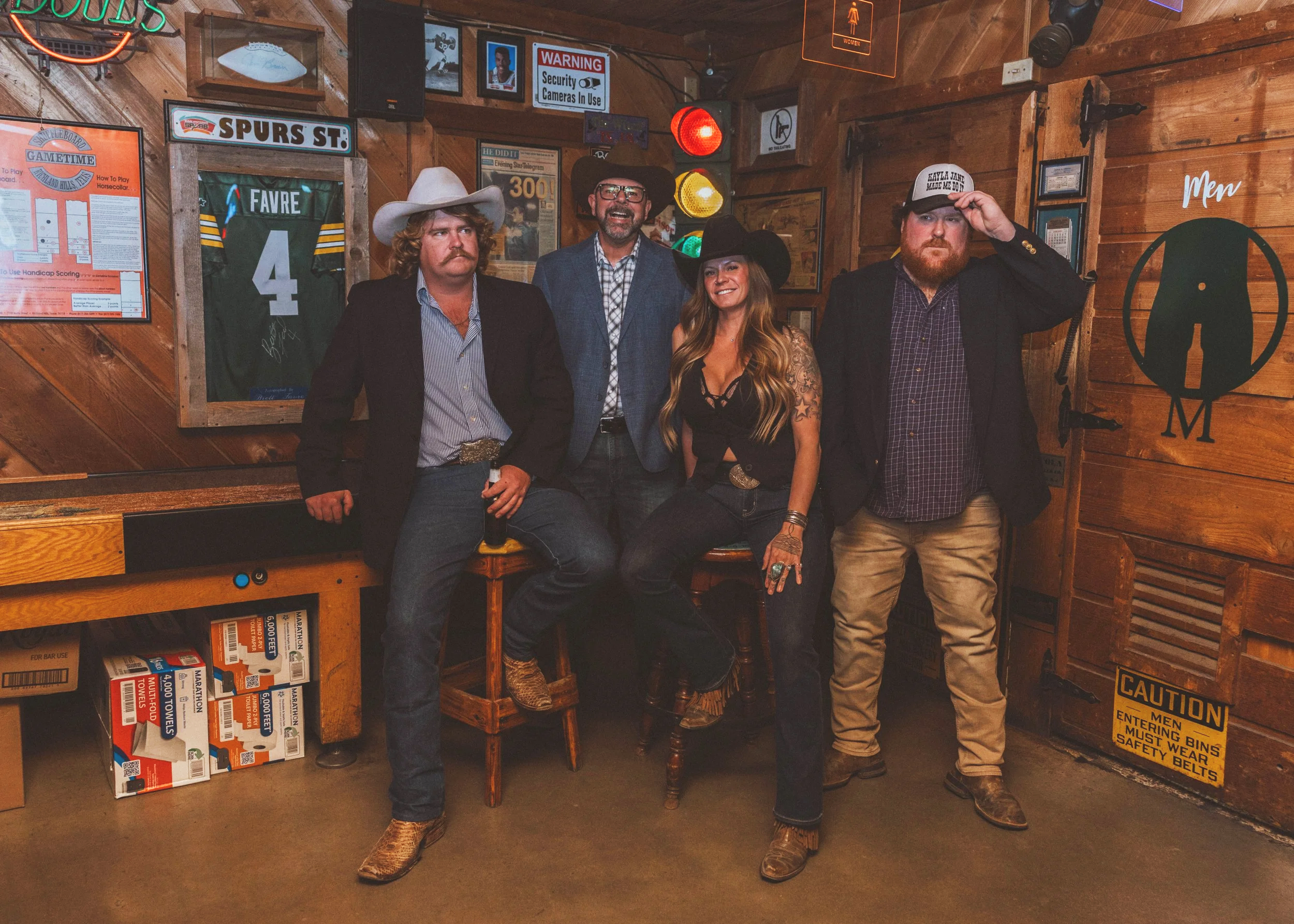 Four people dressed in Western and cowboy attire standing inside a wooden clubhouse or bar decorated with sports memorabilia, signs, and warning notices.
