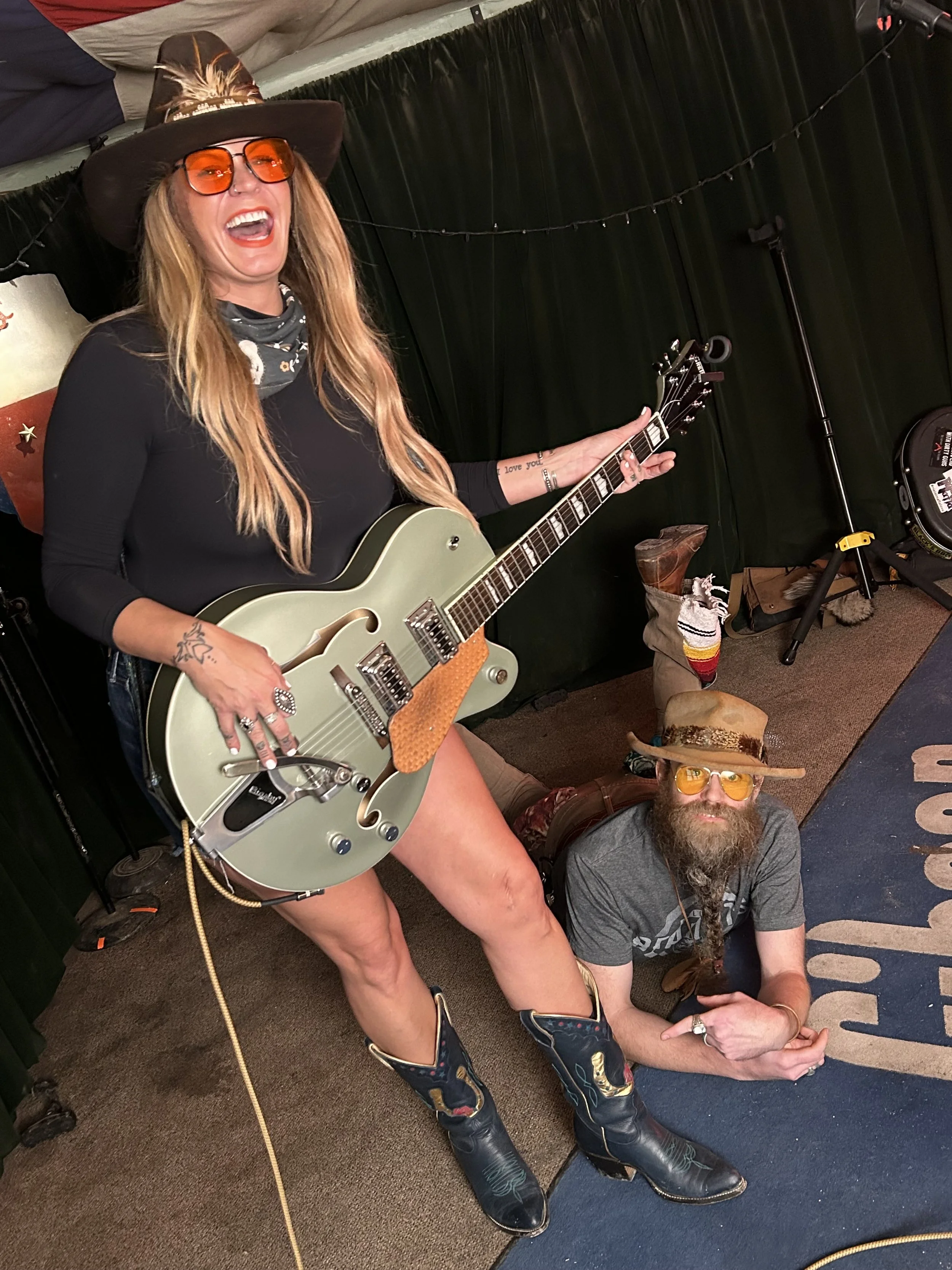 A woman wearing a large black cowboy hat, orange sunglasses, and cowboy boots, holding an electric guitar, standing with a man lying on the floor in a room with black curtain backdrop and string lights. The man has a beard, is wearing a hat, glasses,