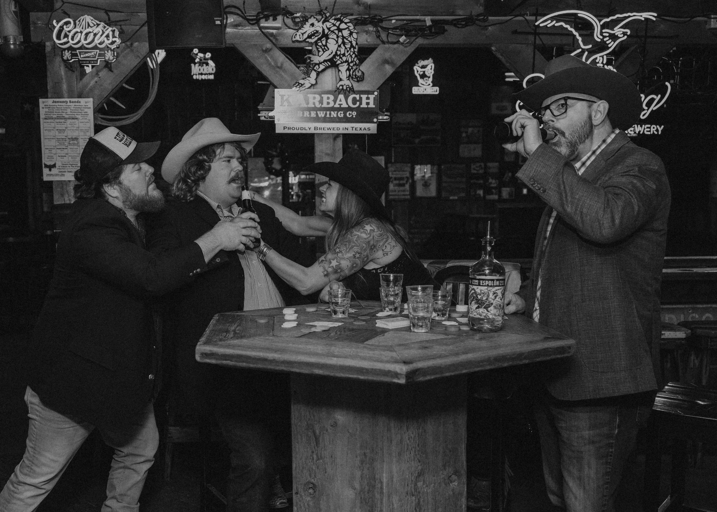 Four people gathered around a wooden table in a bar or pub, engaging in a poker game, with chips and drinks on the table. The background features neon signs, including a Coors sign, a dragon, and a sign for Karbach Brewing Company, with exposed wooden beams and hanging wires, all in black-and-white.