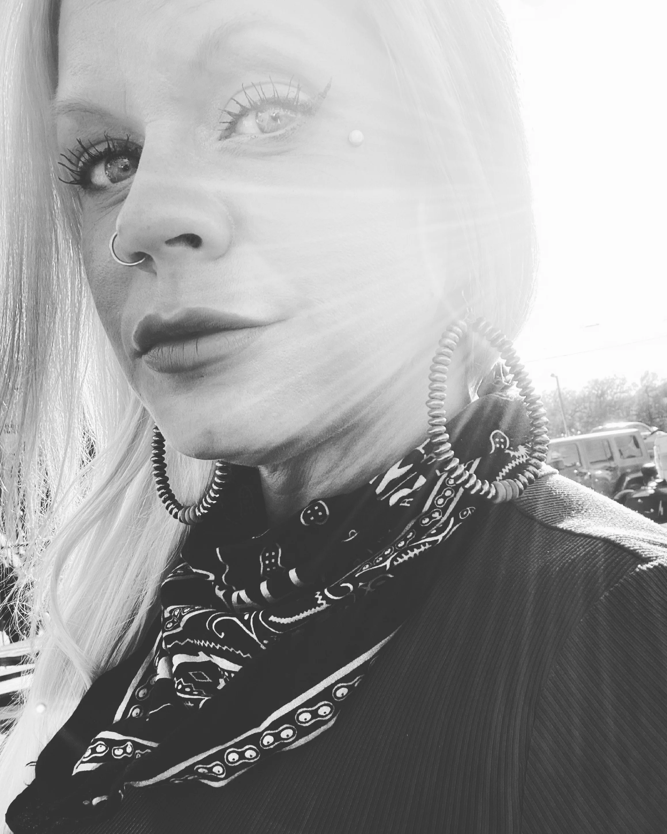 Close-up of a woman with long light hair, wearing hoop earrings, a bandana around her neck, and a jacket, outdoors in sunlight.