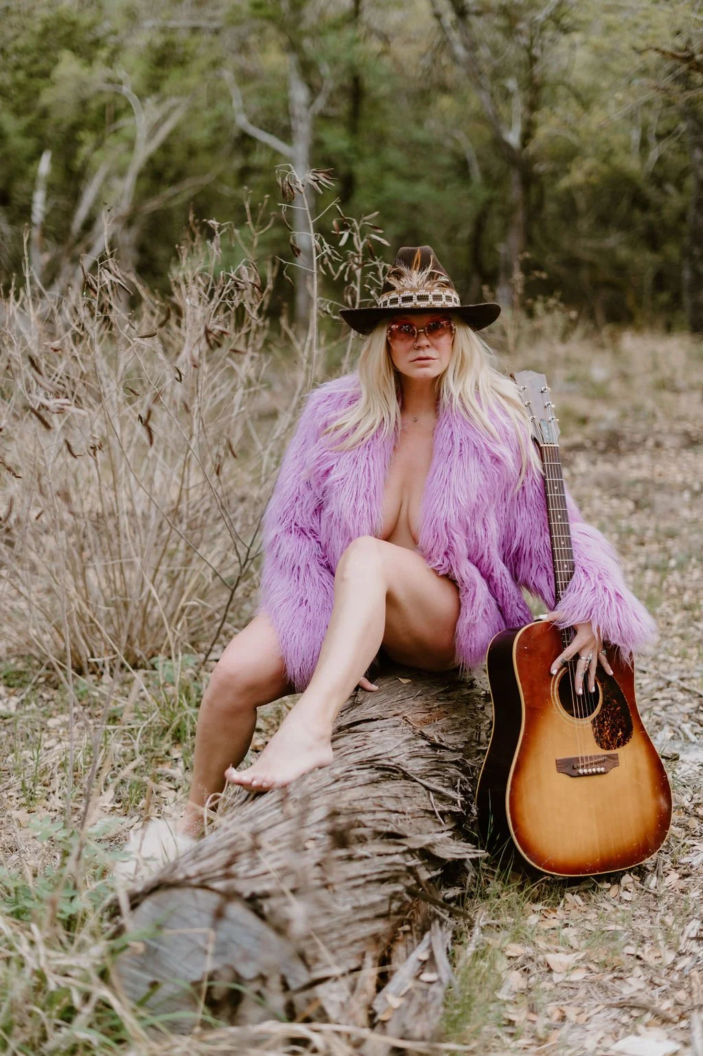 A woman with long blonde hair wearing a purple fur coat, sunglasses, and a feathered hat, sitting on a fallen tree trunk in a wooded outdoor setting, holding an acoustic guitar.