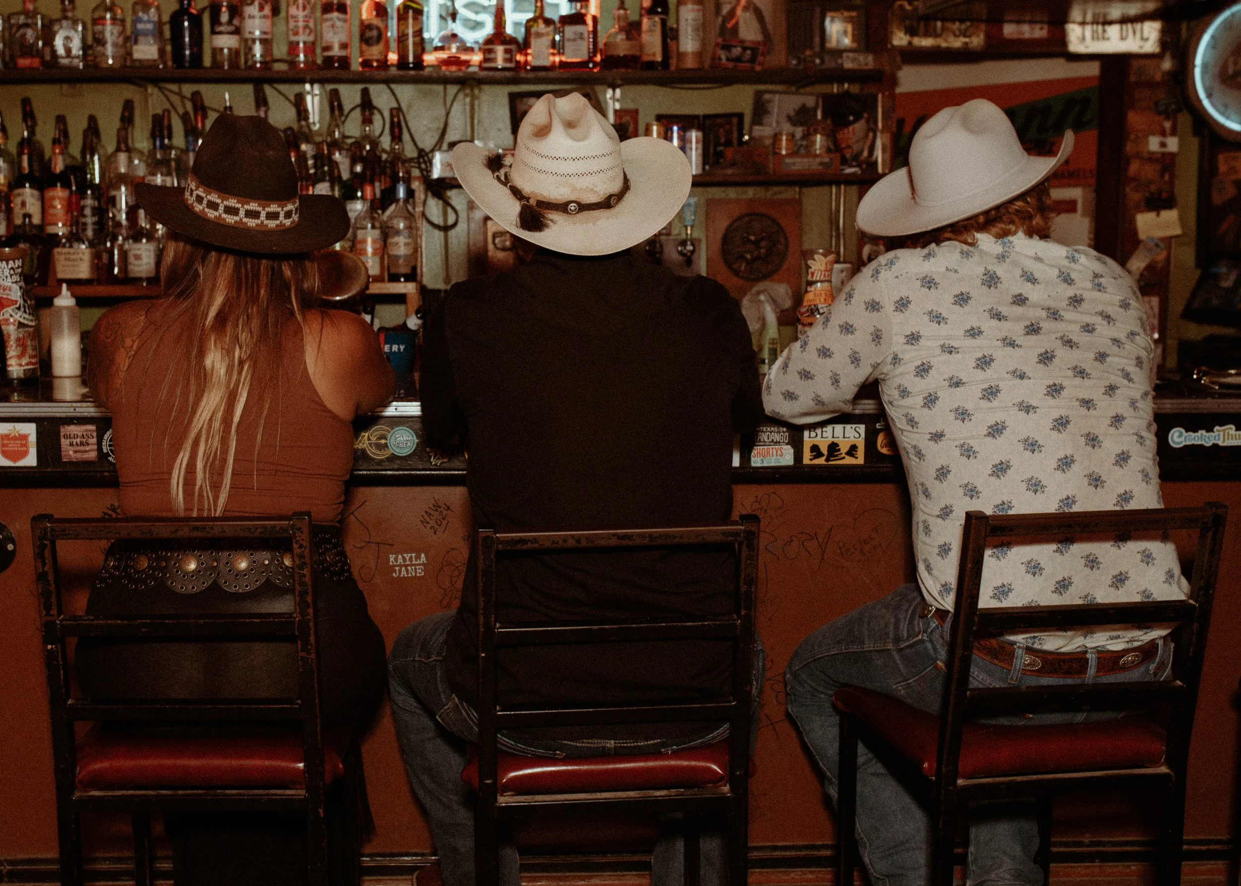 Three people with cowboy hats sitting at a bar with bottles lining the shelves behind them.