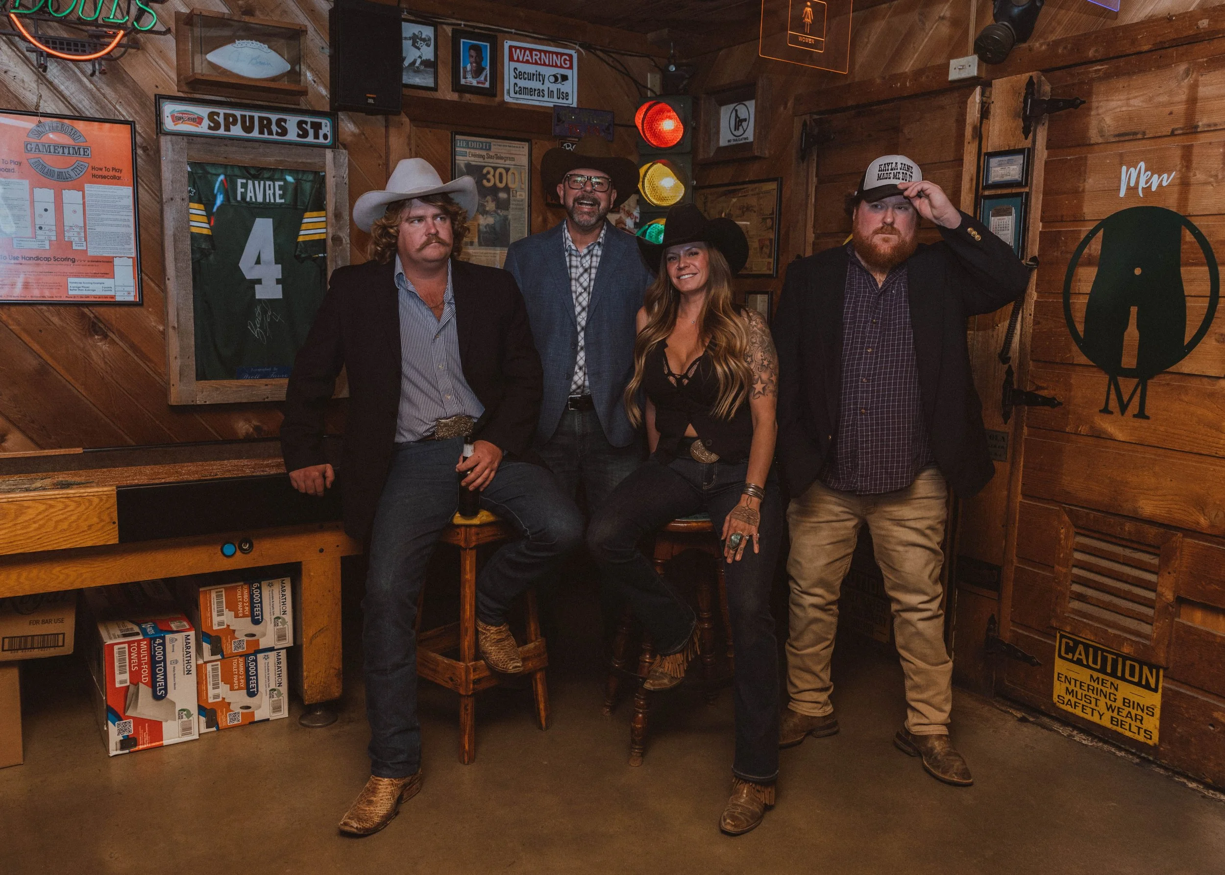 Four people dressed in Western and cowboy attire are posing inside a rustic wooden bar or restaurant. The two men on each end wear cowboy hats; the man on the left has a mustache and patterned shirt, while the man on the right has a beard and is tipp