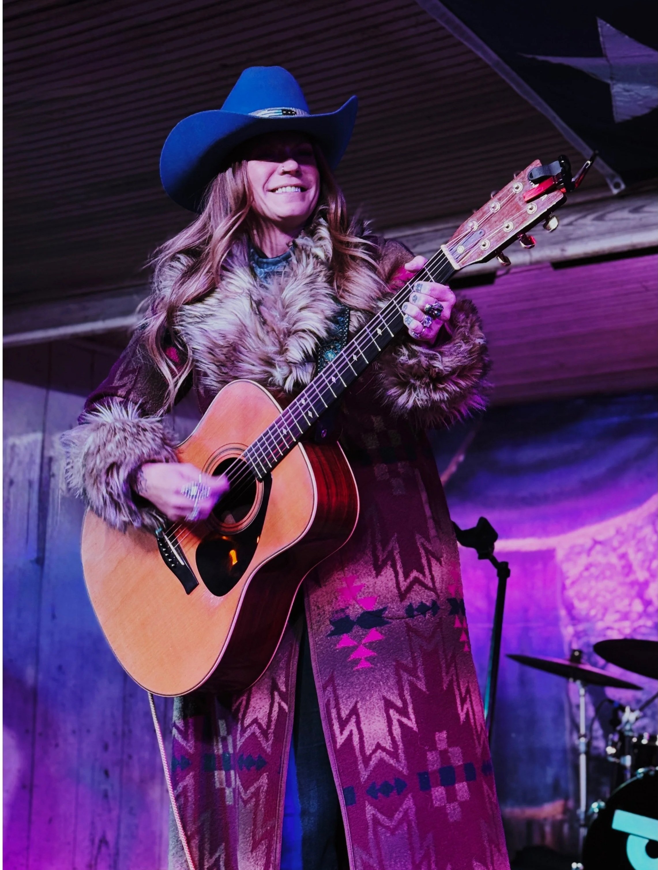 A woman wearing a blue cowboy hat and a fur-lined coat playing an acoustic guitar on a stage with purple lighting.