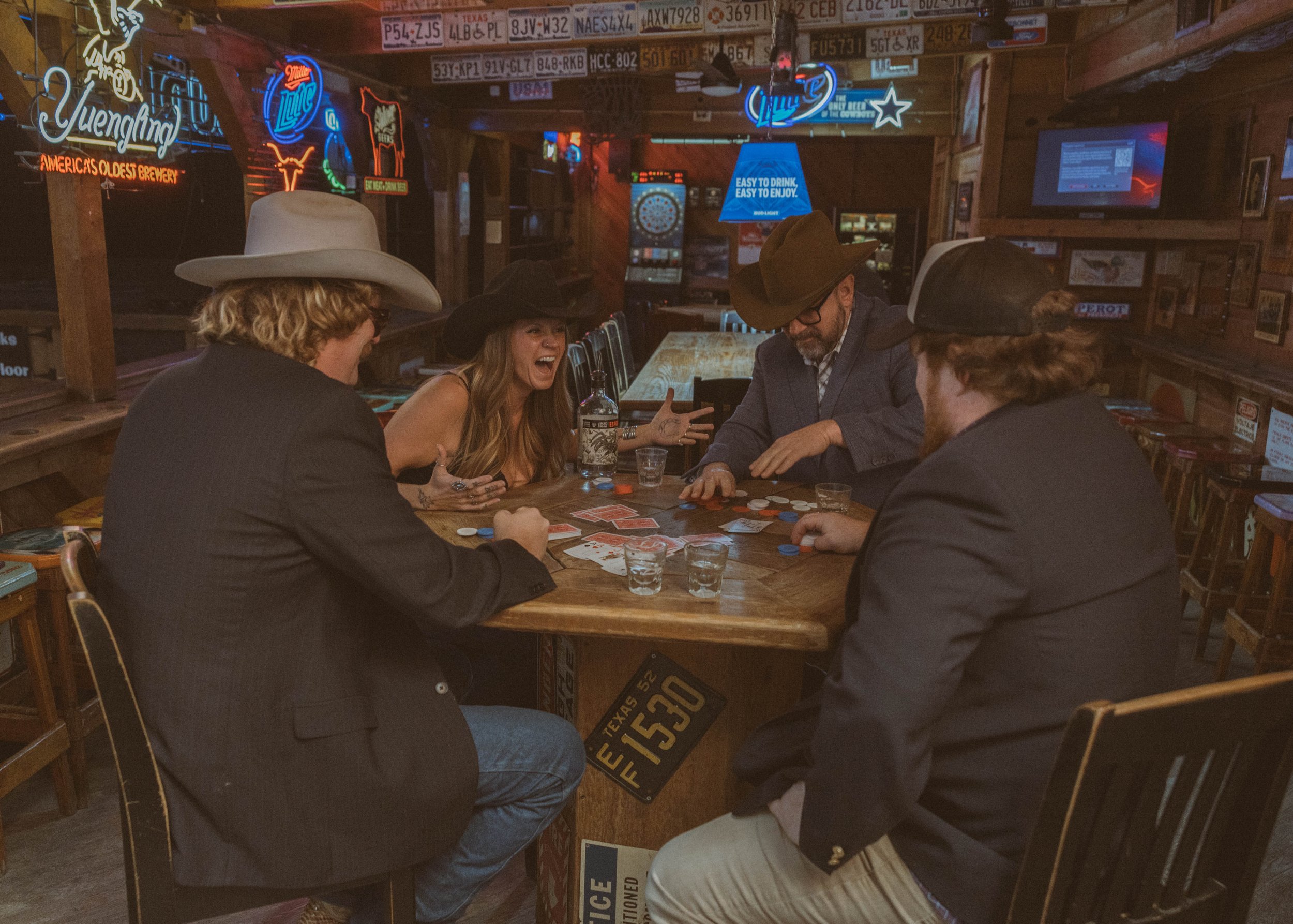Four people in cowboy hats playing poker at a wooden bar in a dimly lit bar with neon signs and hanging license plates.