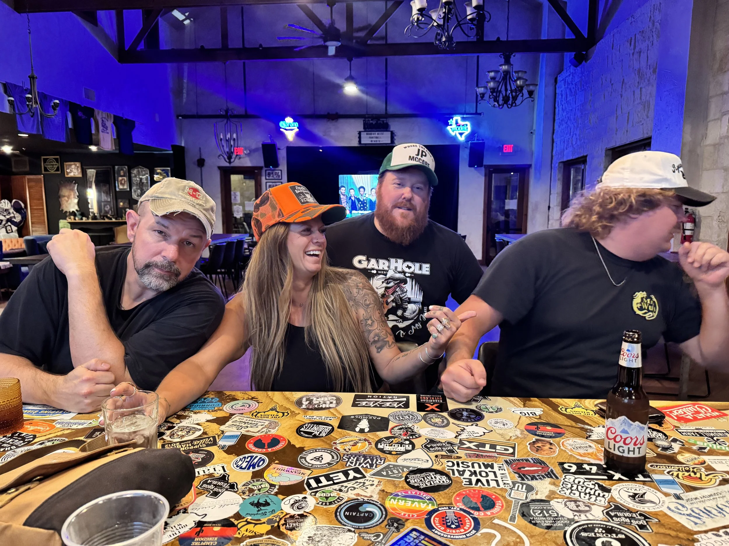A group of four people sitting at a table in a bar or restaurant, wearing caps and smiling. The table is covered with stickers and there are drinking glasses and bottles on it. The background shows neon signs and framed pictures on the wall.