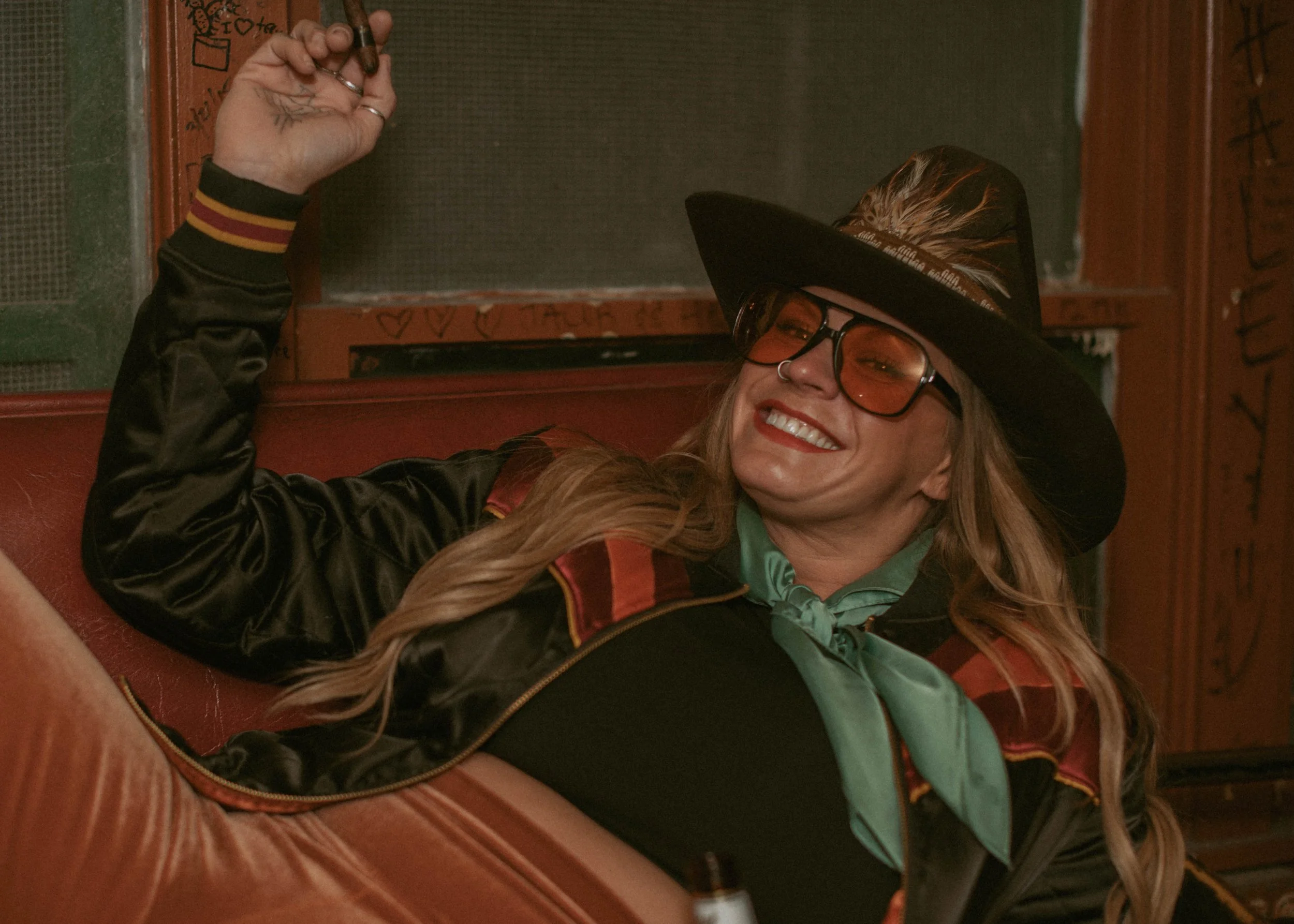 A woman smiling while taking a selfie, wearing large glasses, a black hat with feathers, and a black jacket with a red patch, sitting on a red leather seat in a dimly lit room with wooden walls.