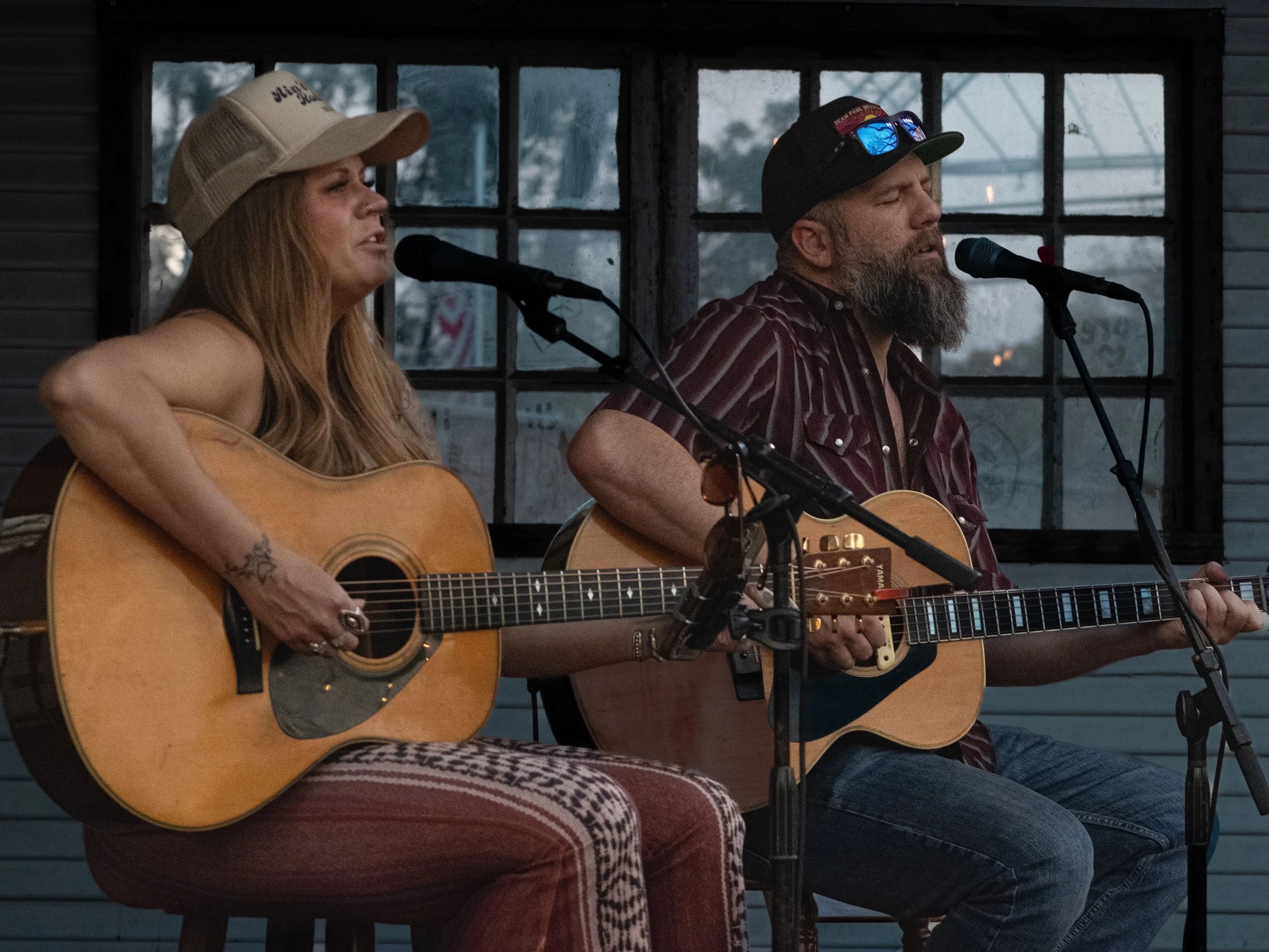 Two musicians playing acoustic guitars and singing into microphones on a stage with a window in the background.