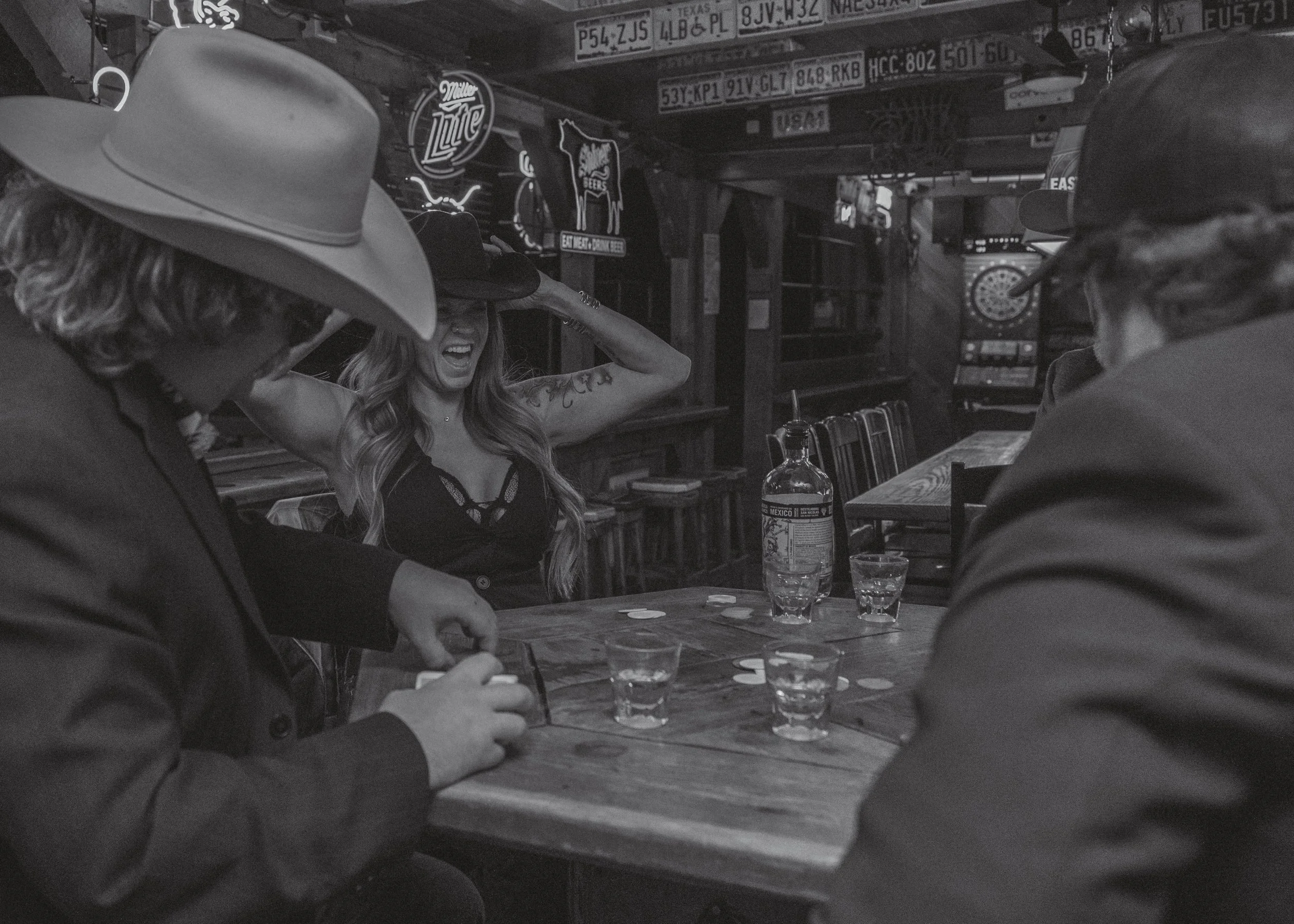 People sitting at a bar or restaurant, engaged in conversation. A woman with long hair and a tattoo on her arm is smiling and holding her head with her hands. There are glasses and bottles on the table. The background has signs with license plates an