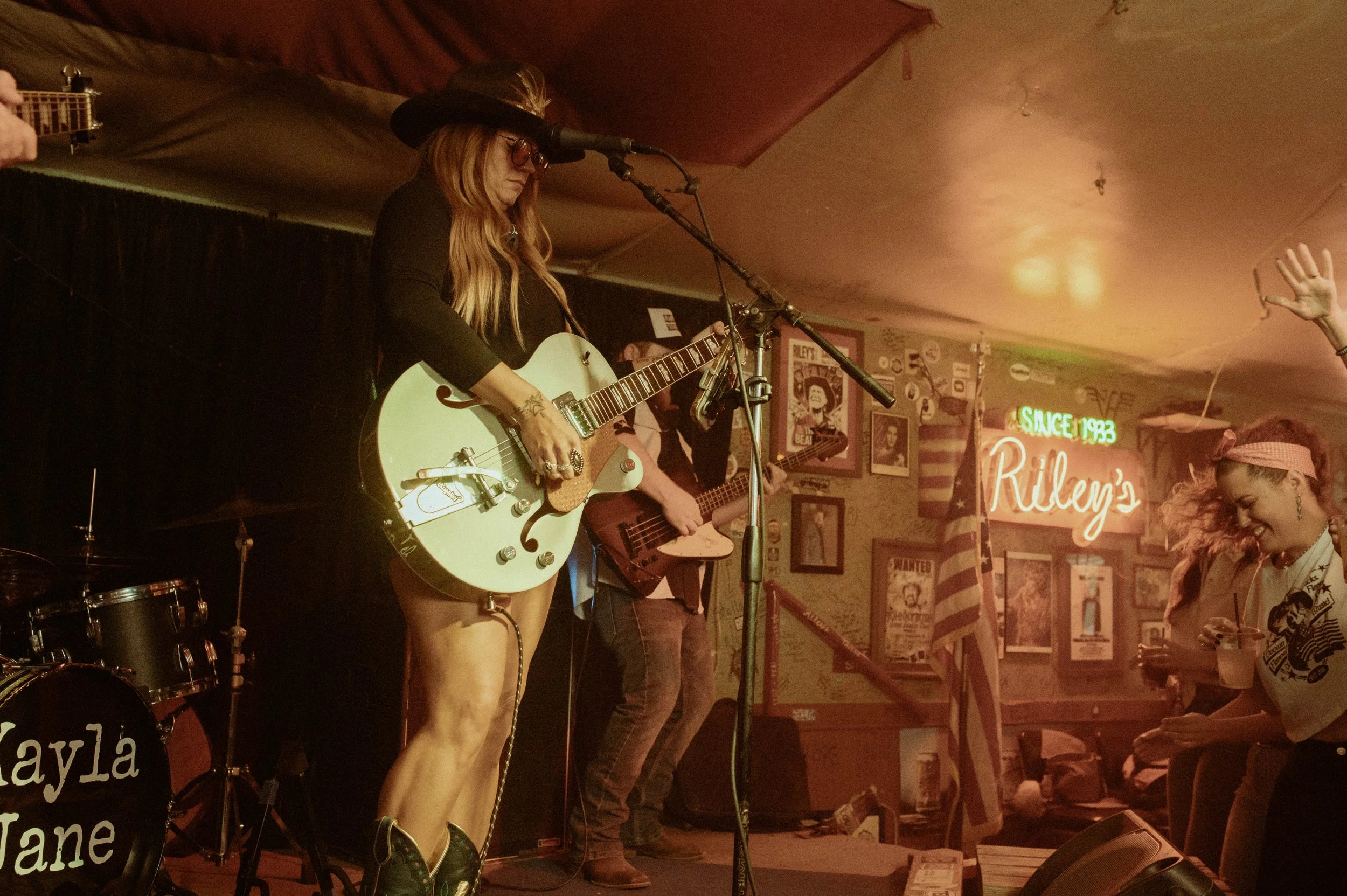 A woman with long hair, wearing glasses, a black hat, and black clothing, playing an electric guitar and singing into a microphone on a stage in a dimly lit bar or music venue.