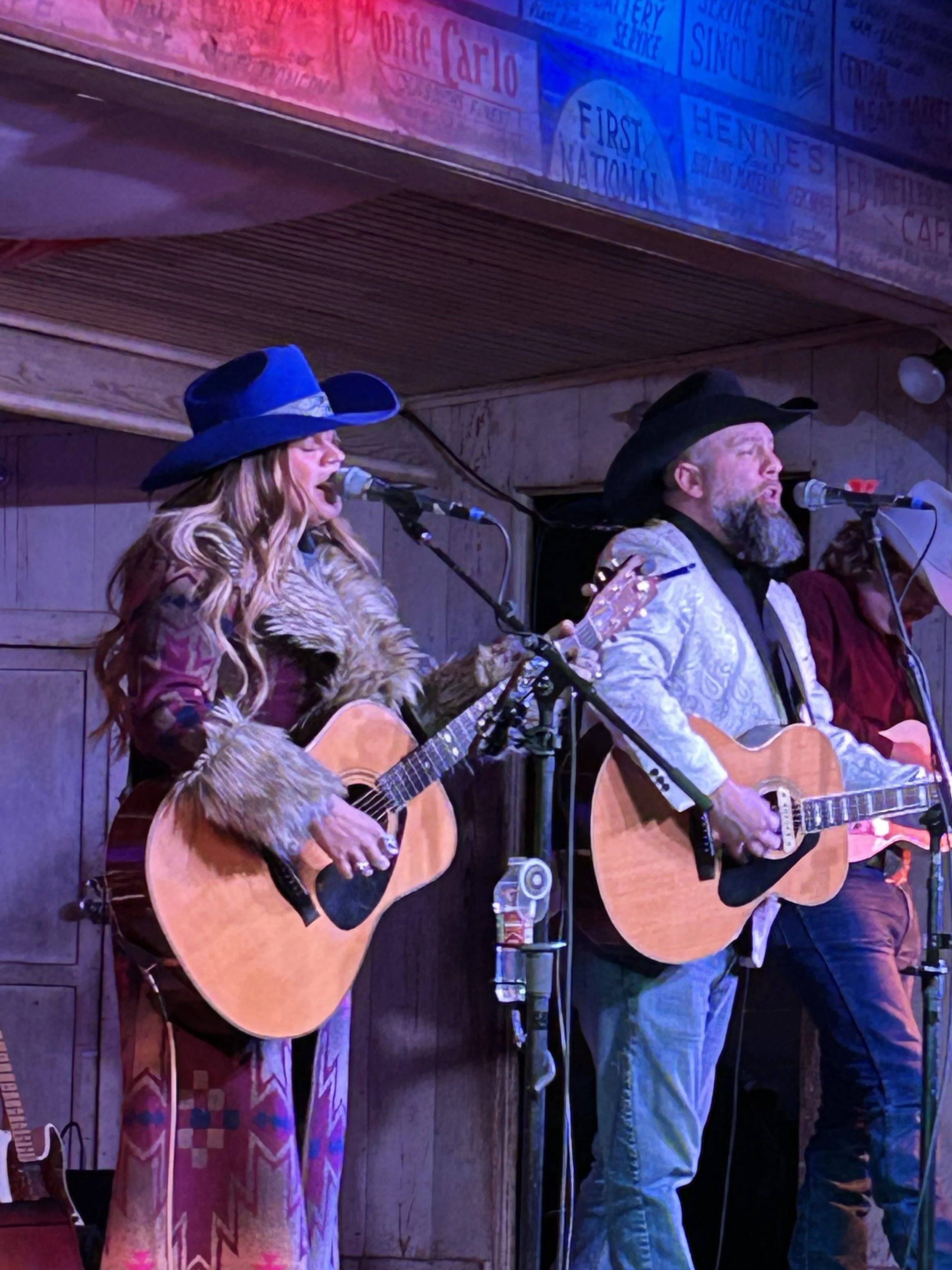 Two musicians playing acoustic guitars and singing into microphones in a dimly lit venue. The woman on the left has long wavy hair, is wearing a large blue hat, a colorful patterned dress, and a furry jacket. The man on the right has a beard, is wearing a black cowboy hat, a light-colored patterned jacket, and a black shirt. There are signs on the wall behind them, and some equipment including a drink on a small table.