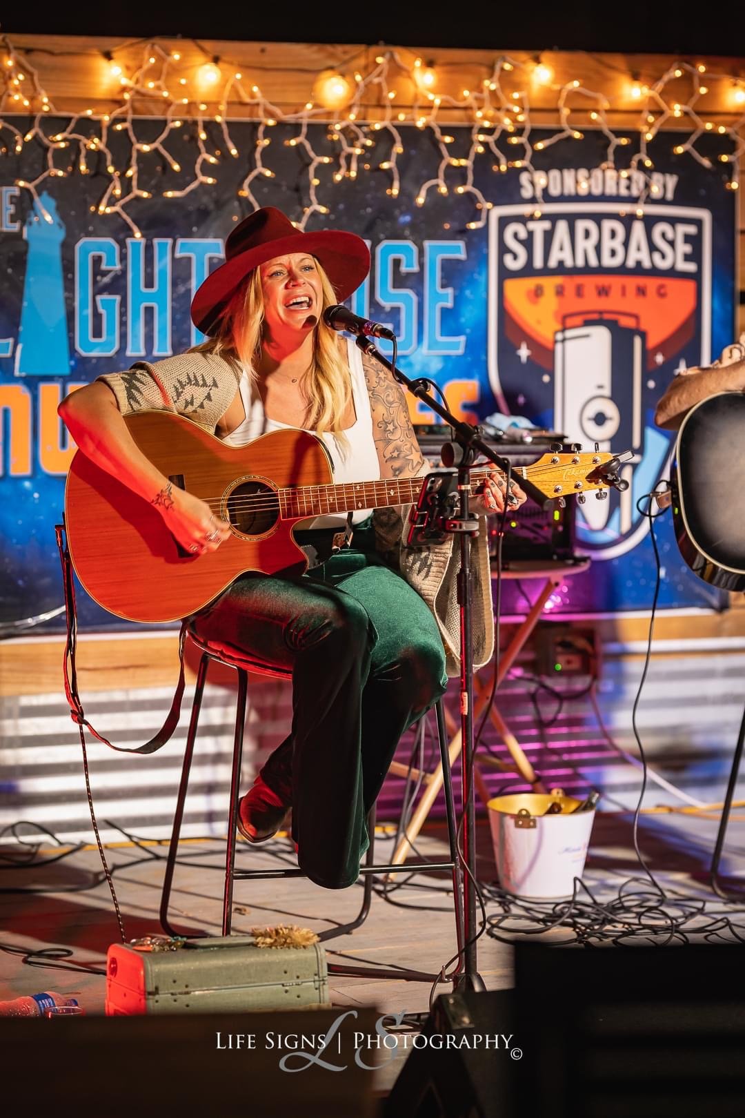 A woman with long blonde hair wearing a wide-brimmed red hat, white tank top, and green pants, singing passionately while playing an acoustic guitar at a stage with string lights and a backdrop featuring sponsorship logos.