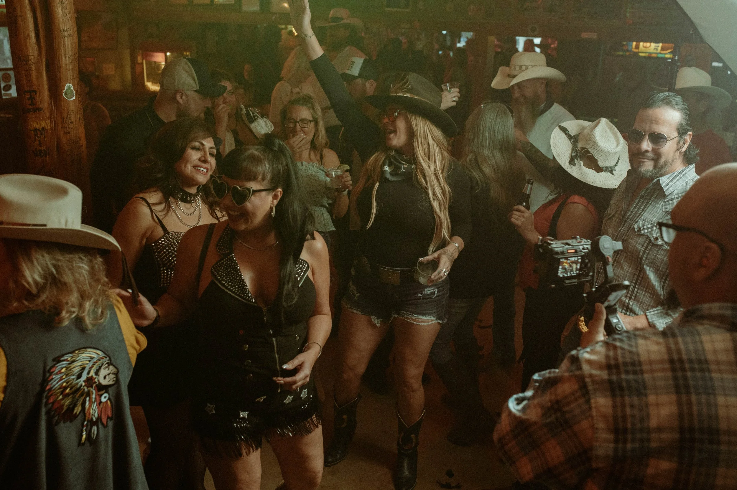 A group of people at a lively party or bar, dressed in Western and country attire, including cowboy hats, sunglasses, and jewelry. They are socializing, holding drinks, and enjoying music in a rustic setting.