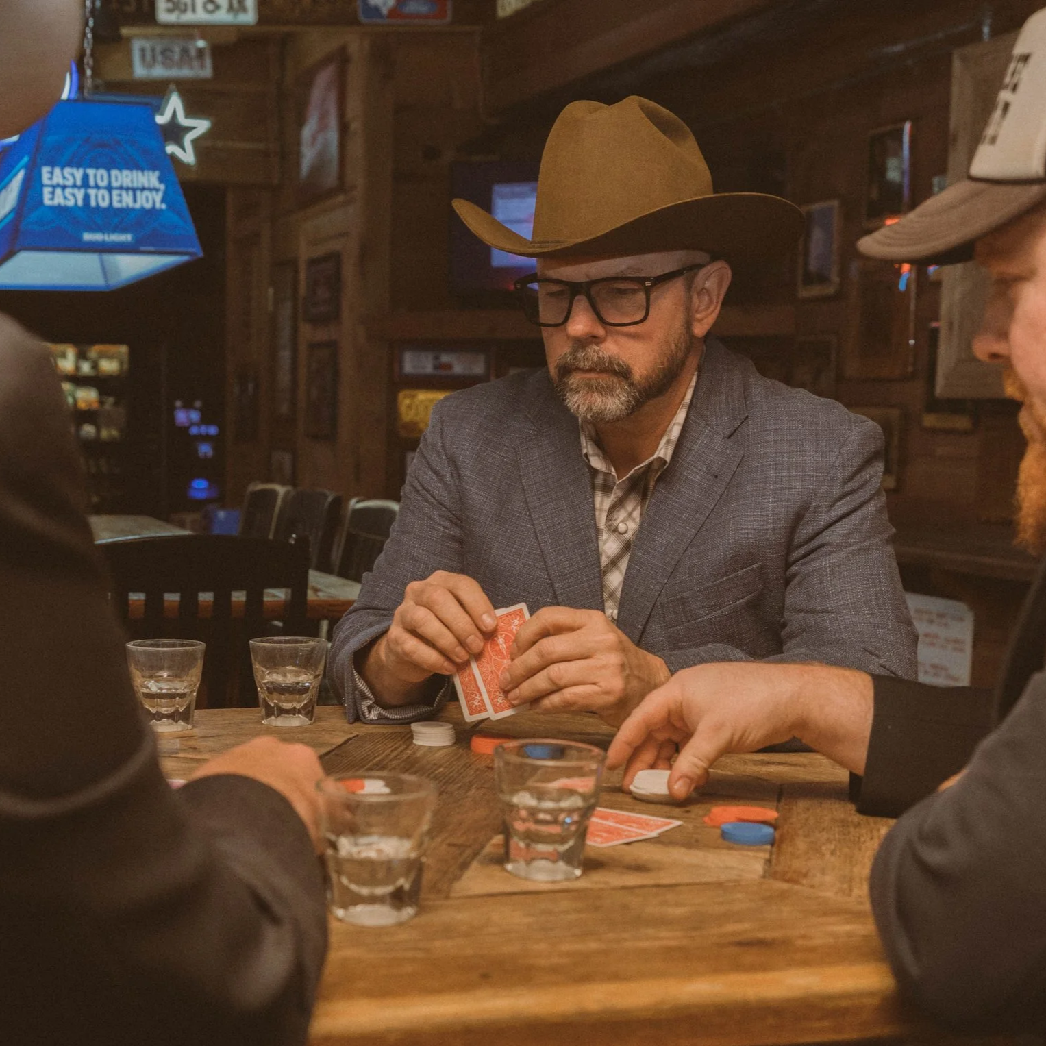 Man wearing cowboy hat, glasses, and plaid shirt holding playing cards at a poker game, with other players and poker chips on a wooden table in a dimly lit bar or restaurant.