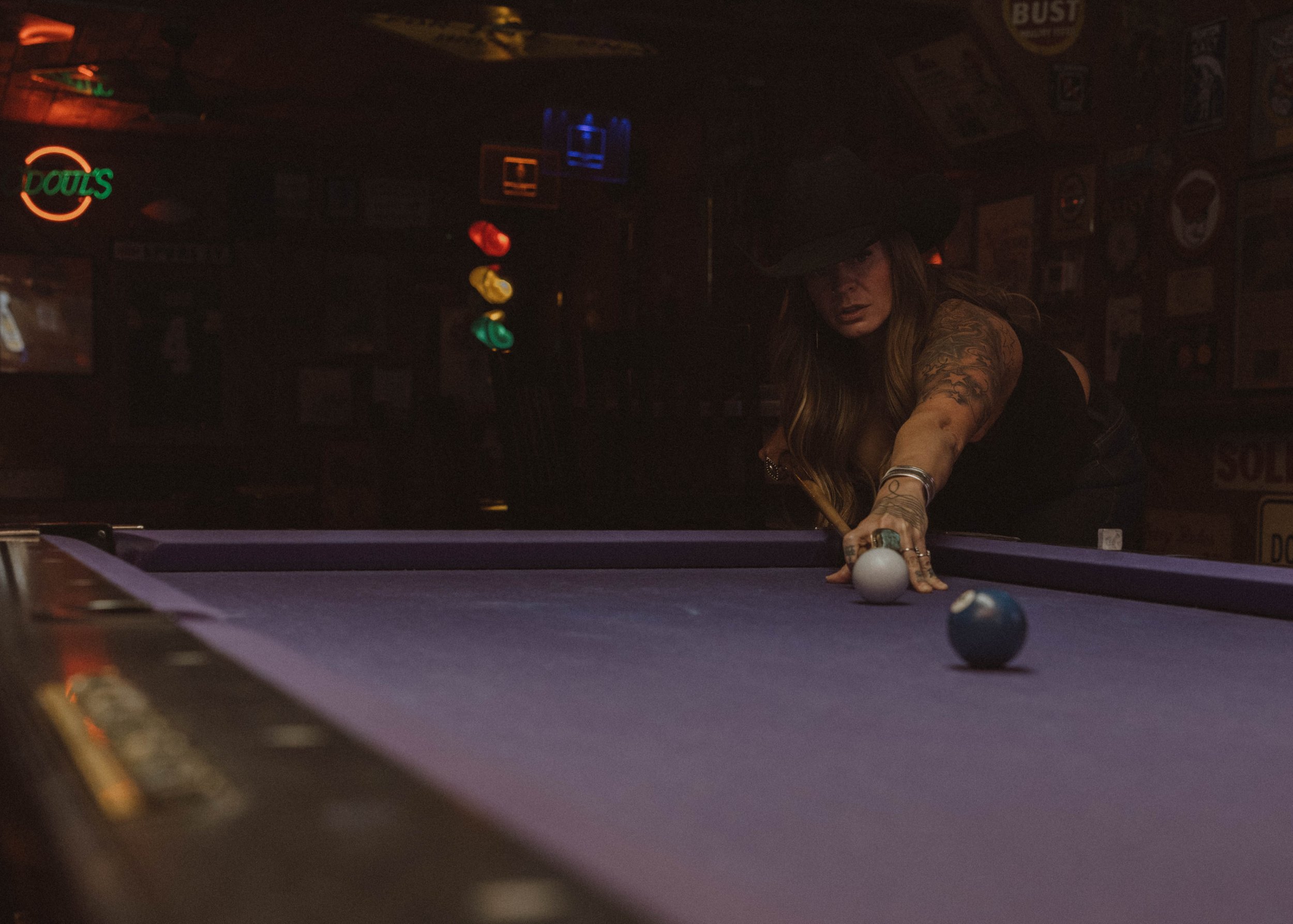 A woman with long hair and tattoos playing pool, leaning over the cue ball, in a dimly lit bar decorated with neon signs and posters.