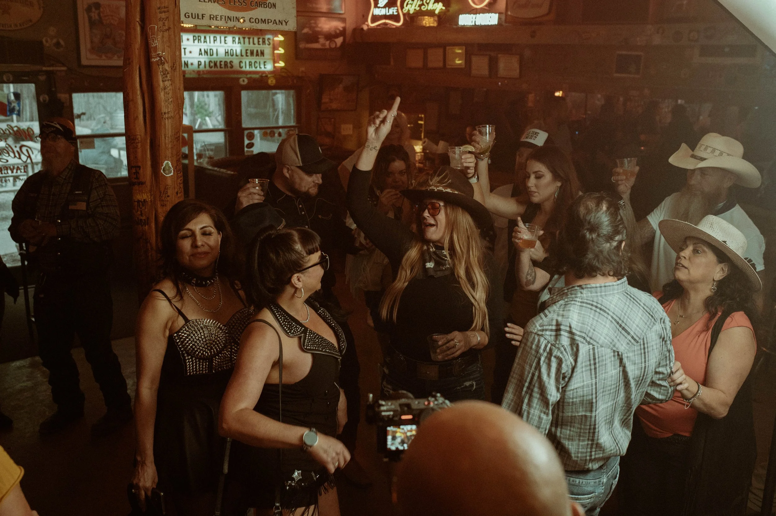 A lively gathering of people in a bar or restaurant, many holding drinks, with neon signs and wooden decor on the walls. Some women are wearing cowboy hats and western-style clothing, suggesting a country or western theme at the event.