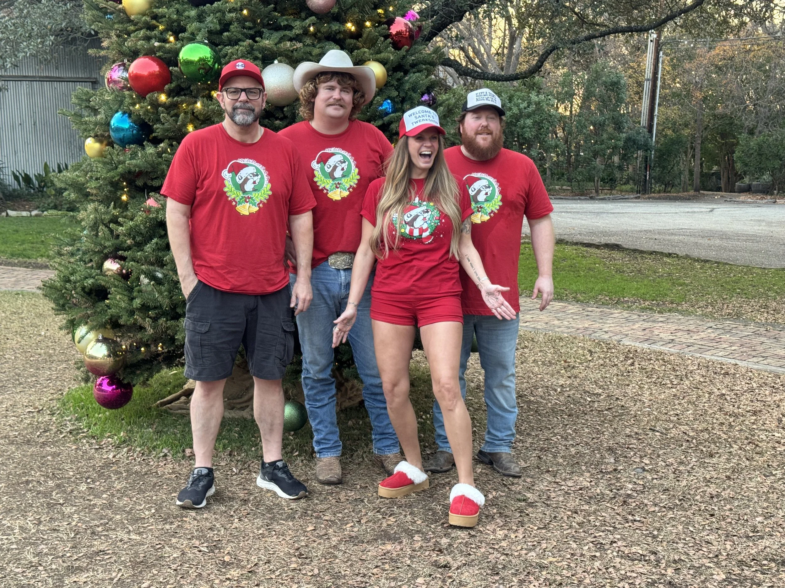 Four people standing outdoors in front of a decorated Christmas tree on a grassy and paved area. They are wearing red Christmas-themed shirts, and two of them have hats. One person has a white cowboy hat, another has a red cap with white text, and th