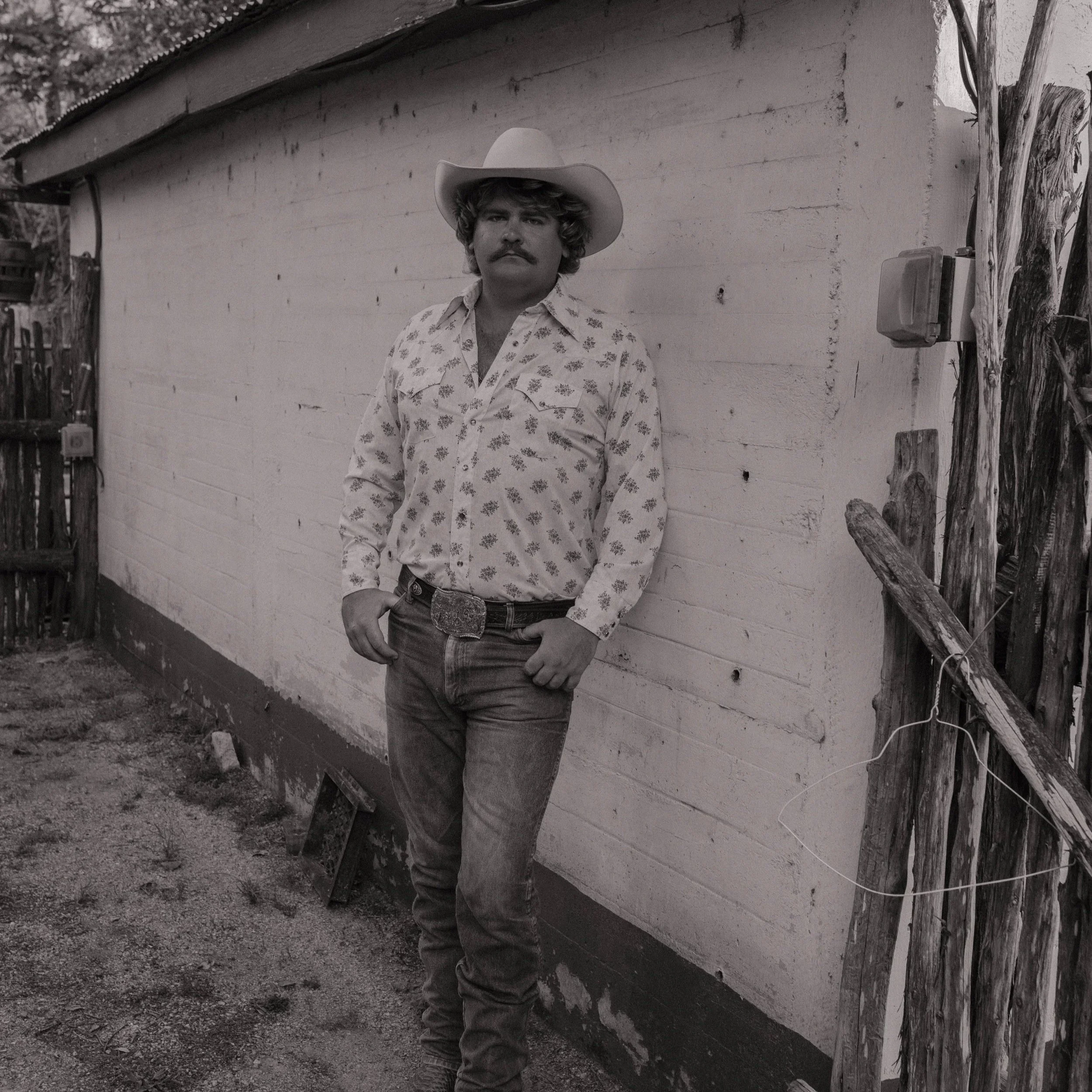 A man with a mustache and curly hair, wearing a cowboy hat and a patterned shirt, stands outdoors next to a wooden building with with a tree in the background.