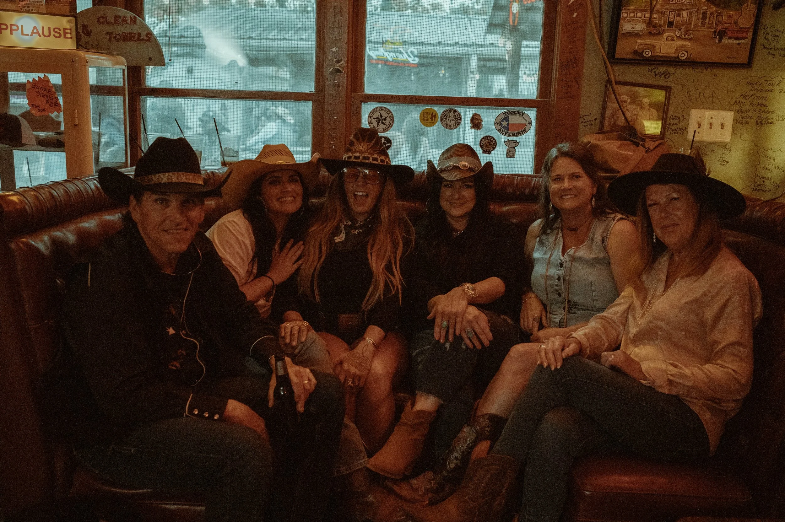 Group of six women sitting on a leather bench inside a restaurant or cafe, wearing cowboy hats, smiling, and posing for a photo.