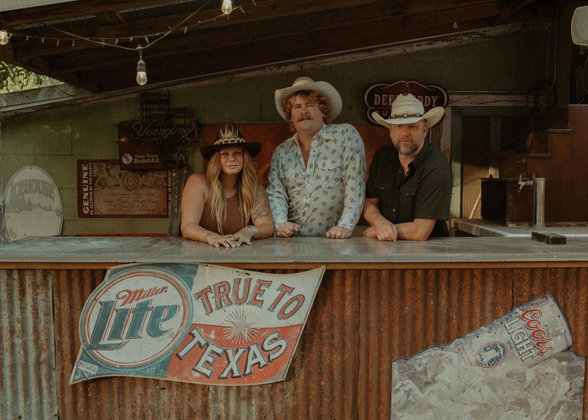 Three people wearing cowboy hats and western clothing standing behind a bar counter at a rustic Texan bar with vintage signs and posters on the wall.