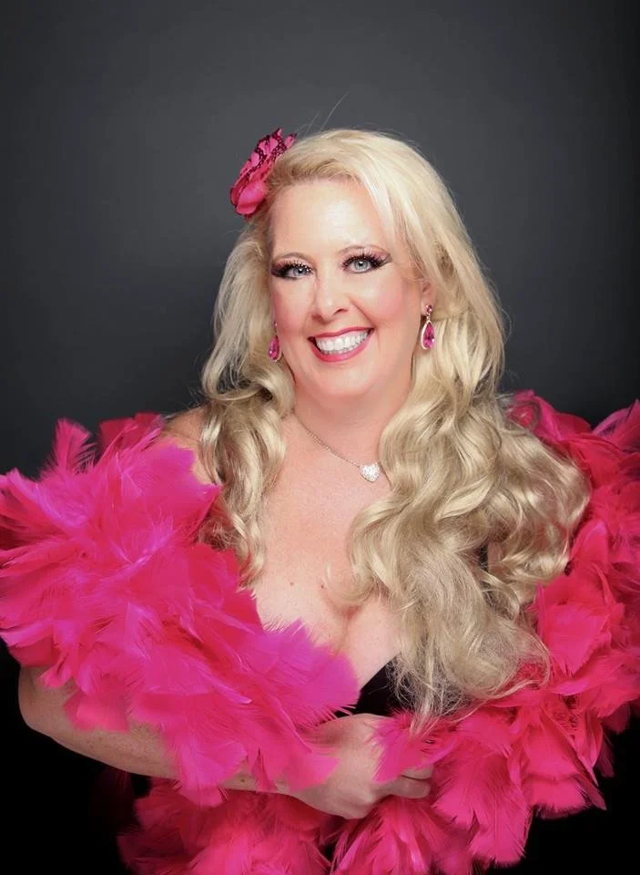 A woman with long, curly blonde hair wearing pink jewelry and a fluffy, pink feather boa, smiling at the camera against a dark gray background.