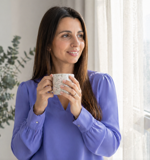 Woman with long brown hair wearing a blue blouse, holding a white speckled mug, standing near a window with light curtains, looking outside.