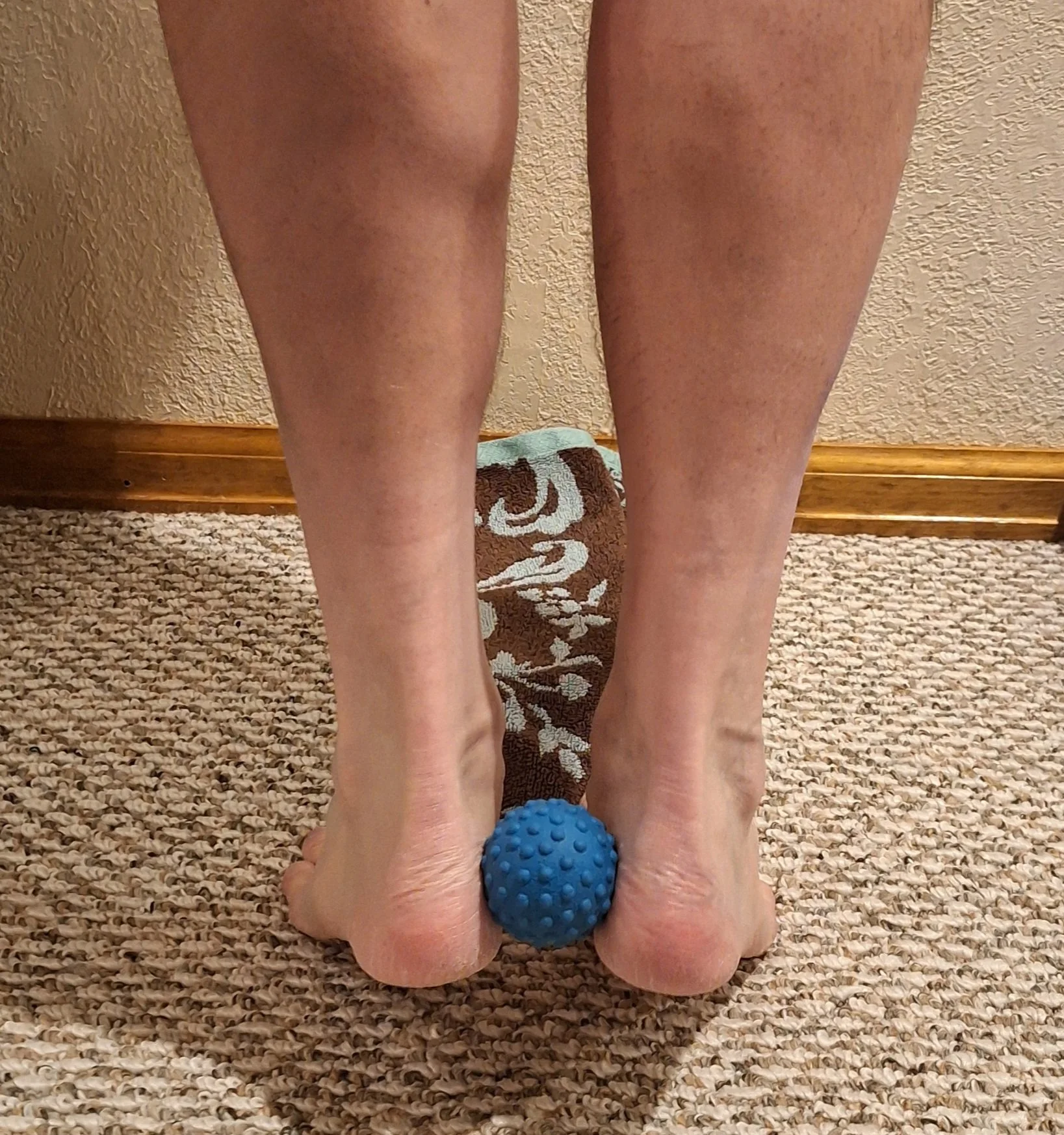 Person kneeling on a carpeted floor, using a blue textured massage ball under their feet for foot therapy.