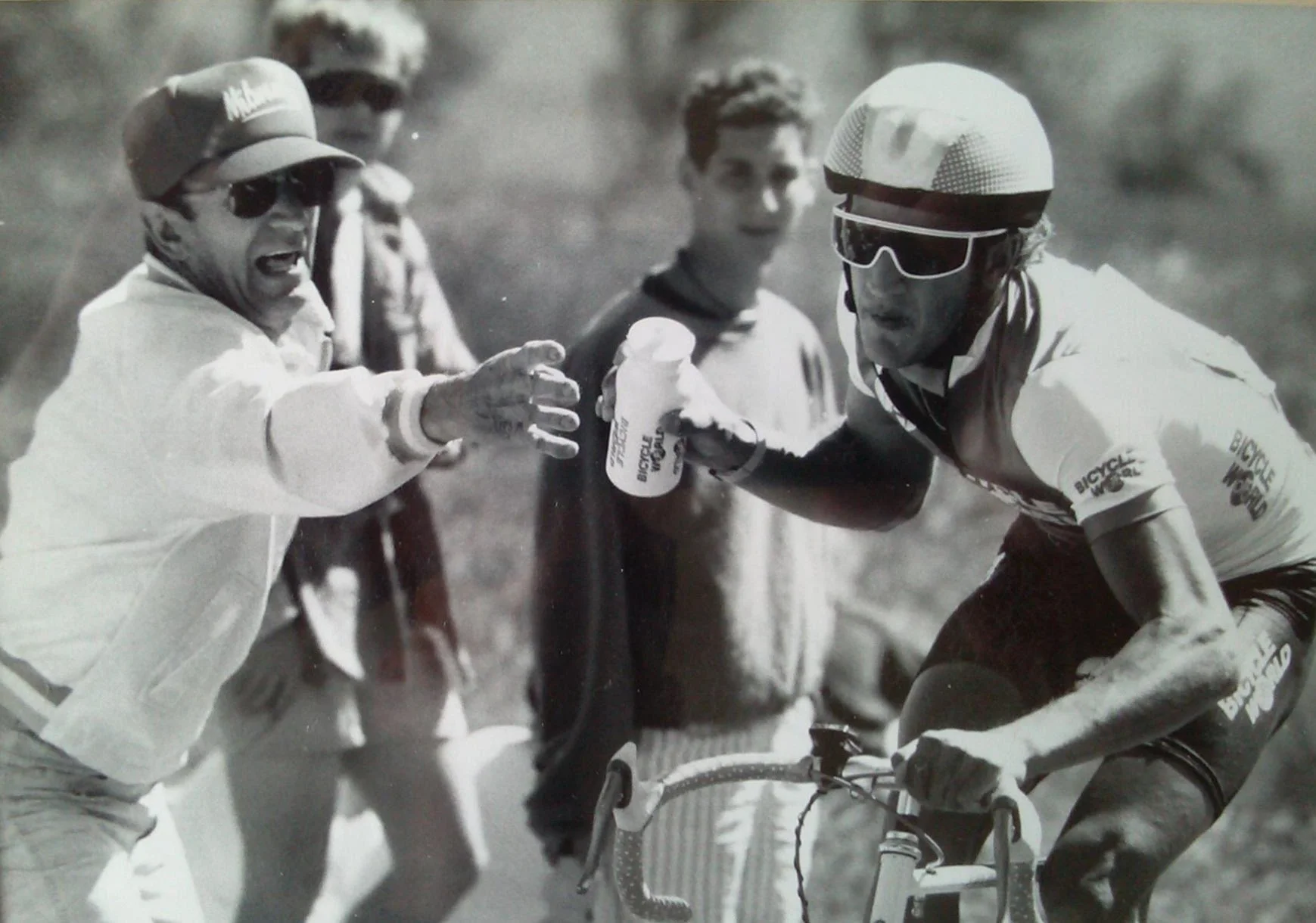 A cyclist in a racing outfit and helmet leans forward on his bike while another person offers him a water bottle during a race or training. Three other individuals stand in the background.