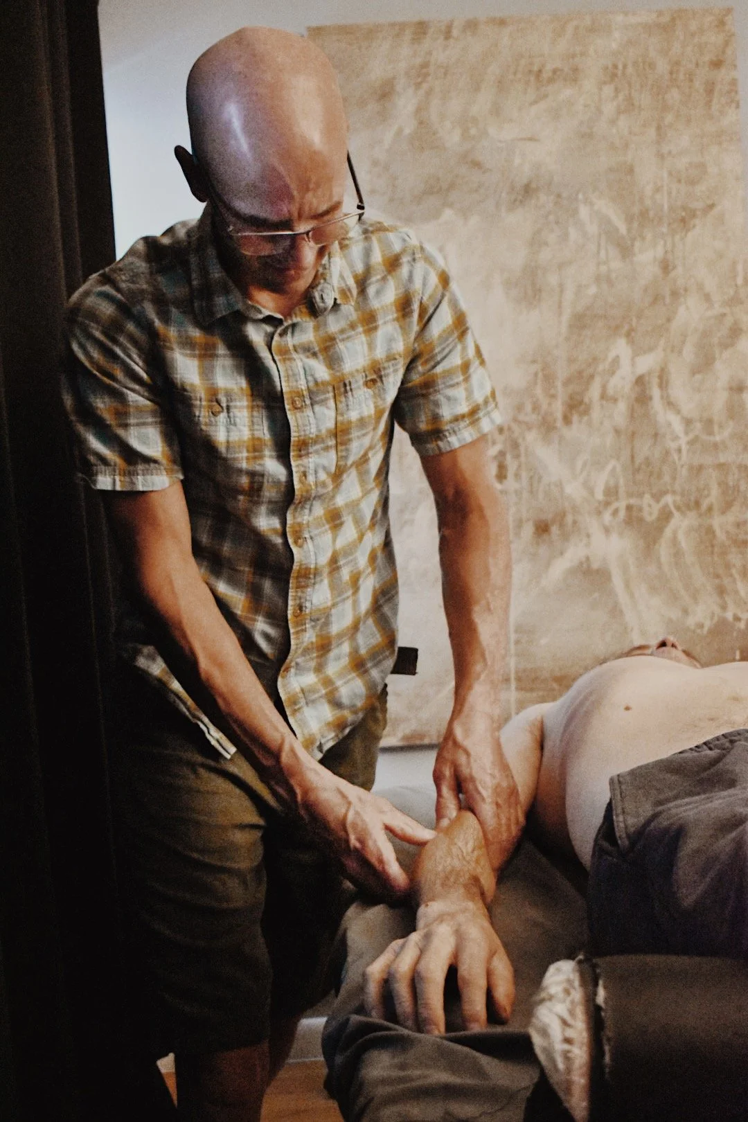 A man giving a massage to another person lying face down on a bed or massage table, in a room with a beige wall and abstract art.