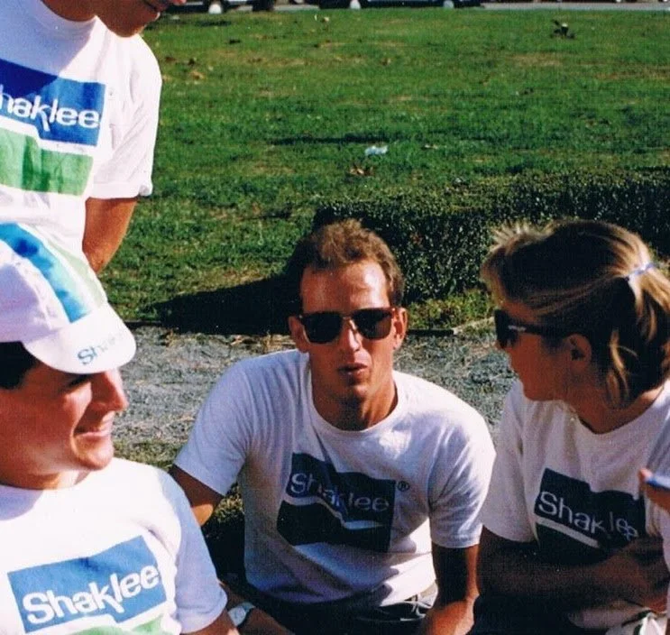 Group of four young adults wearing white Shaklee shirts, sitting and standing outdoors on grass near a stream.