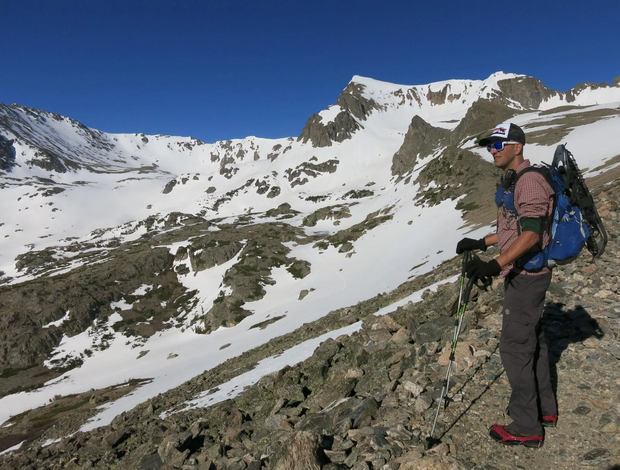 A man hiking in a snowy mountainous landscape wearing sunglasses, a cap, a backpack, and using trekking poles.