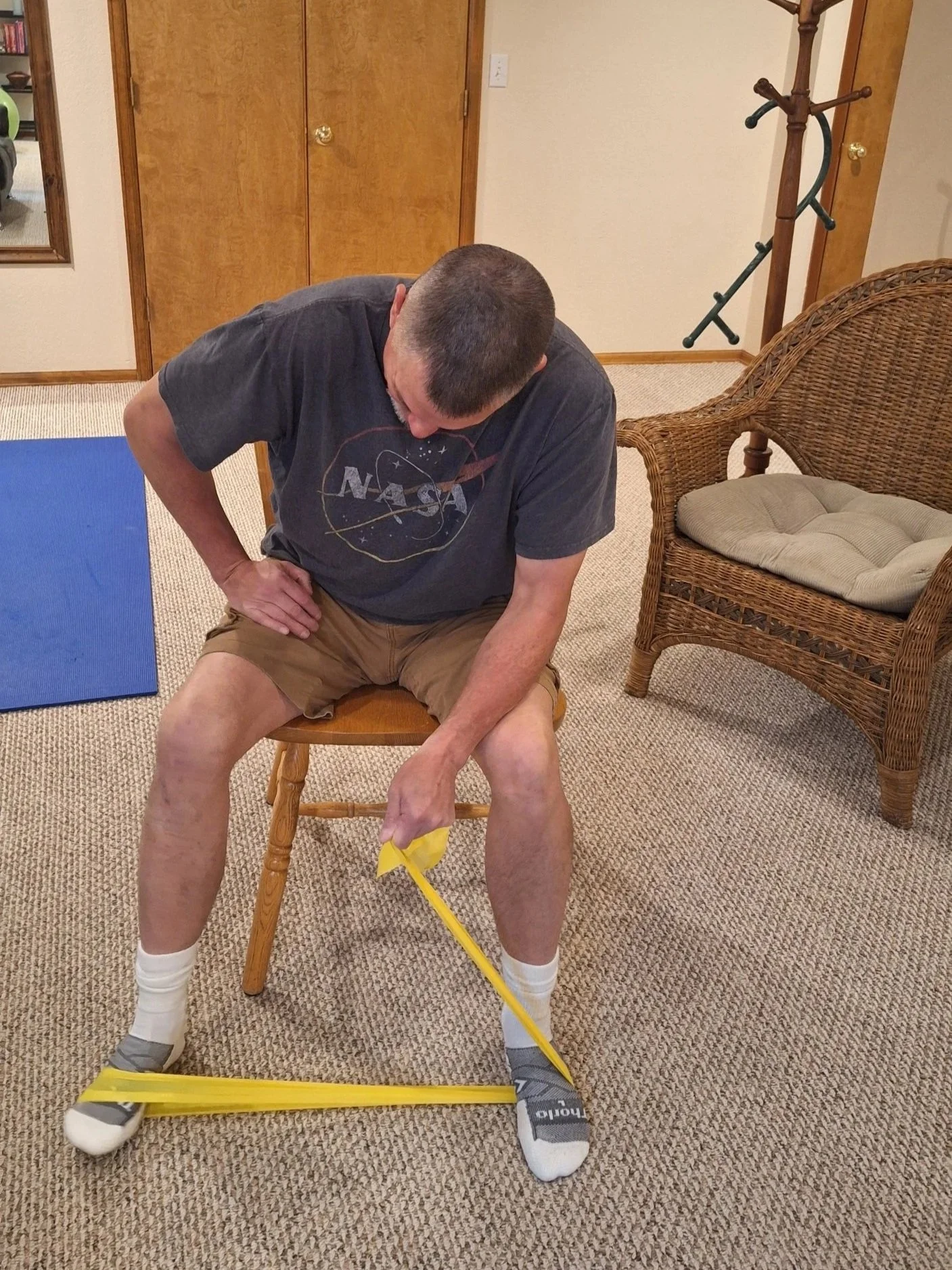 A man with a short haircut, wearing a NASA T-shirt, brown shorts, and socks with slippers, sitting on a wooden chair in a room with beige carpet, holding a yellow object with a long yellow pole attached to his foot, stretching his leg while looking down.