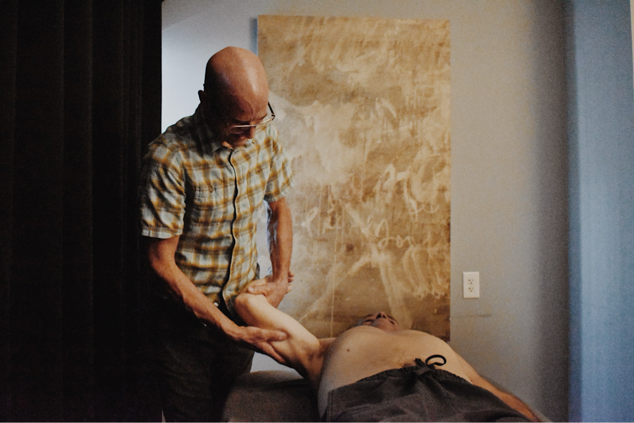 A man giving a massage to another man lying on a massage table in a room with a beige and gray wall, and an electrical outlet visible in the background.