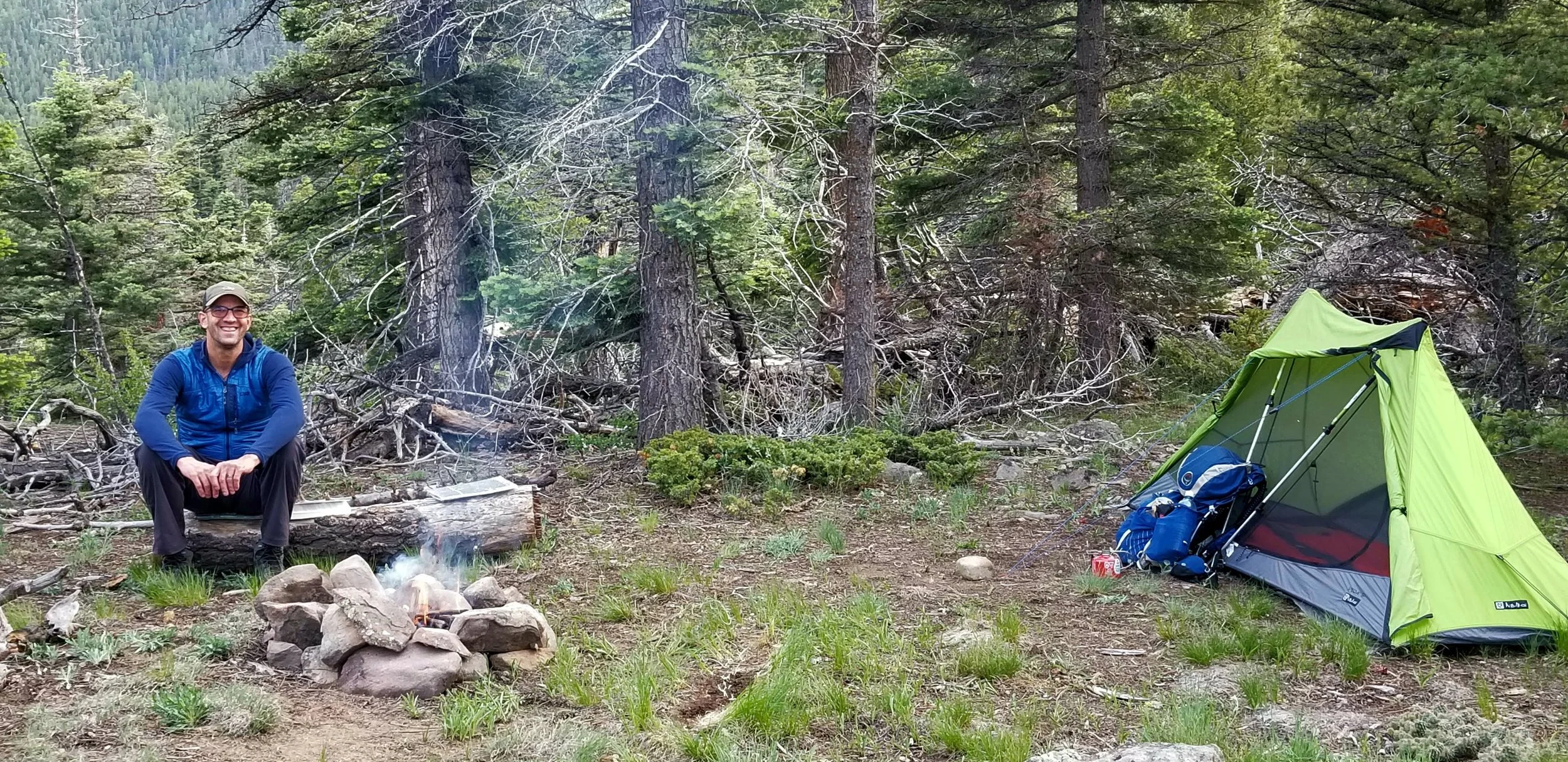 A man smiling and sitting near a campfire in a forest with trees, and a bright green camping tent to the right.