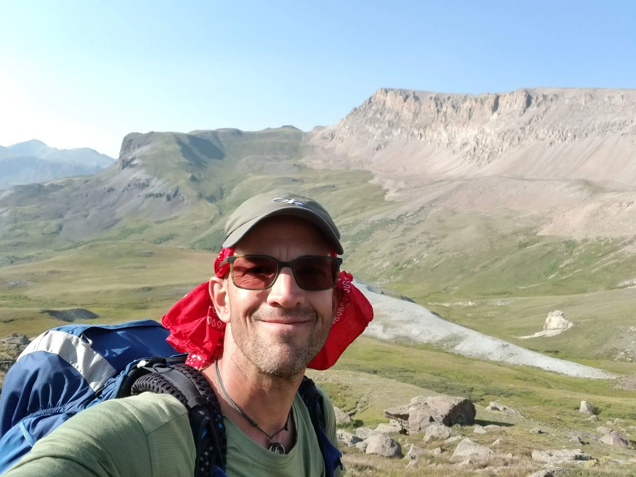 A man with sunglasses and a cap taking a selfie in a mountainous landscape with rolling hills, rocky terrain, and large rock formations under a clear blue sky.