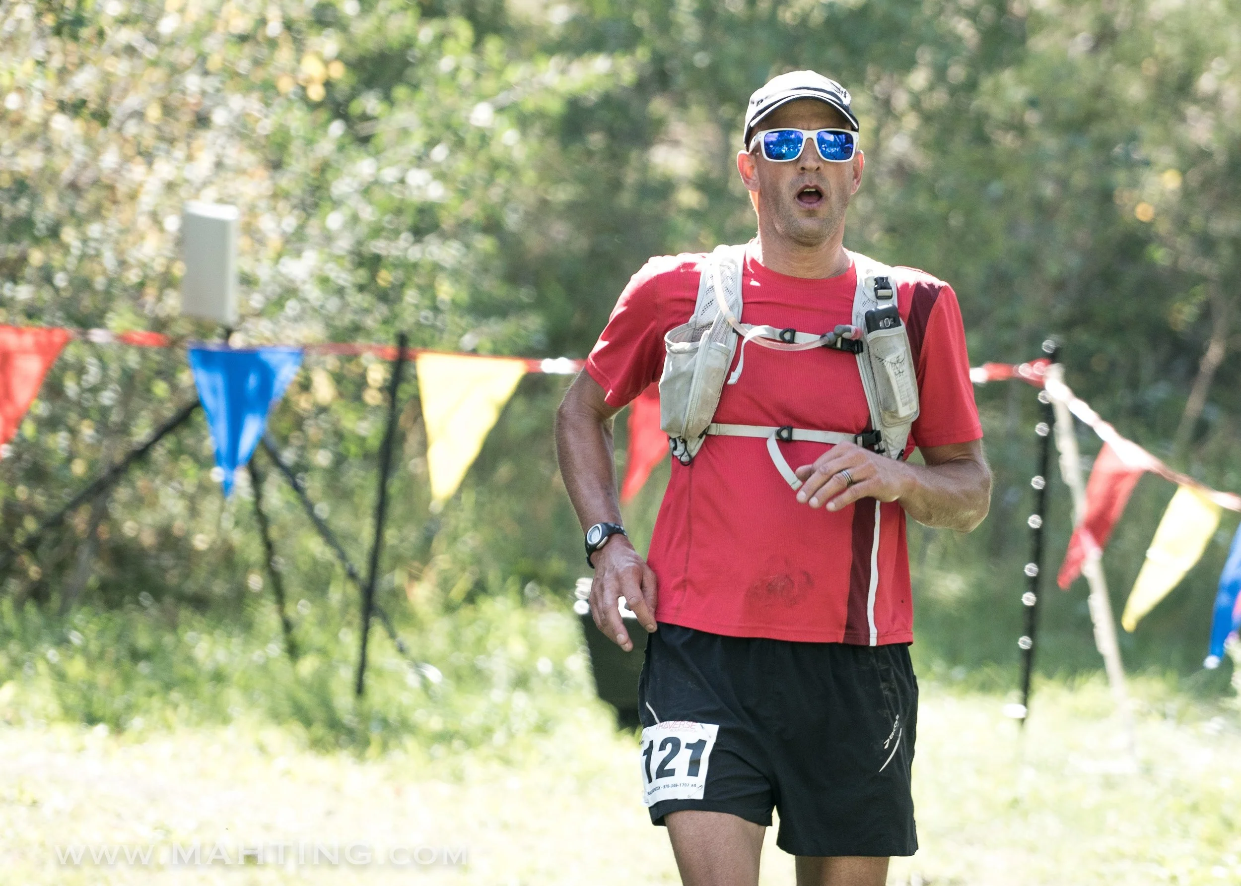 A man in a red athletic shirt and black shorts running outdoors, wearing sunglasses, a hat, a hydration pack, and a race bib number 121, with colorful flags in the background.