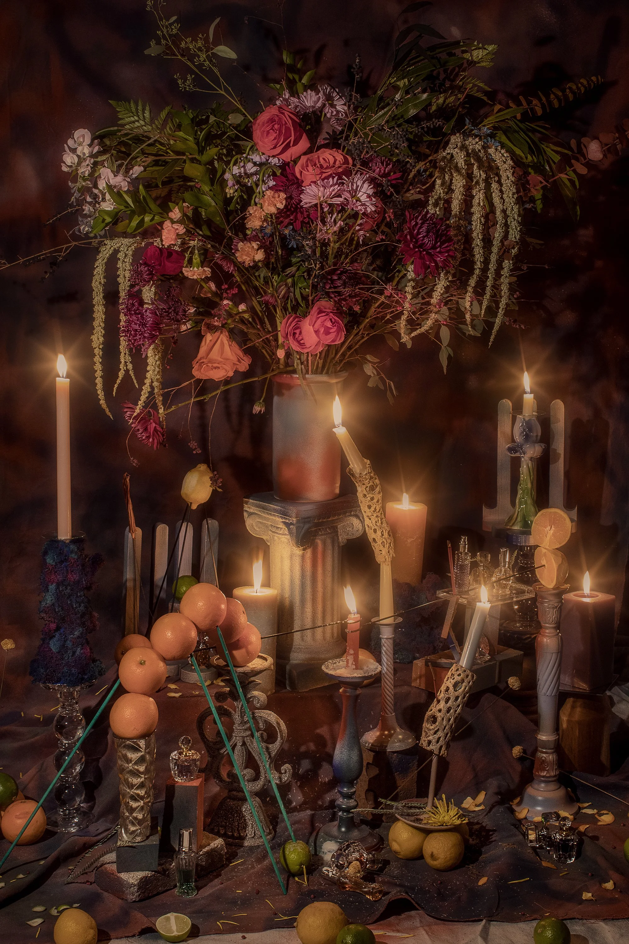 A large bouquet of various flowers in a pinkish-orange vase, surrounded by lit candles, limes, lemons, and decorative items on a table with a dark background.