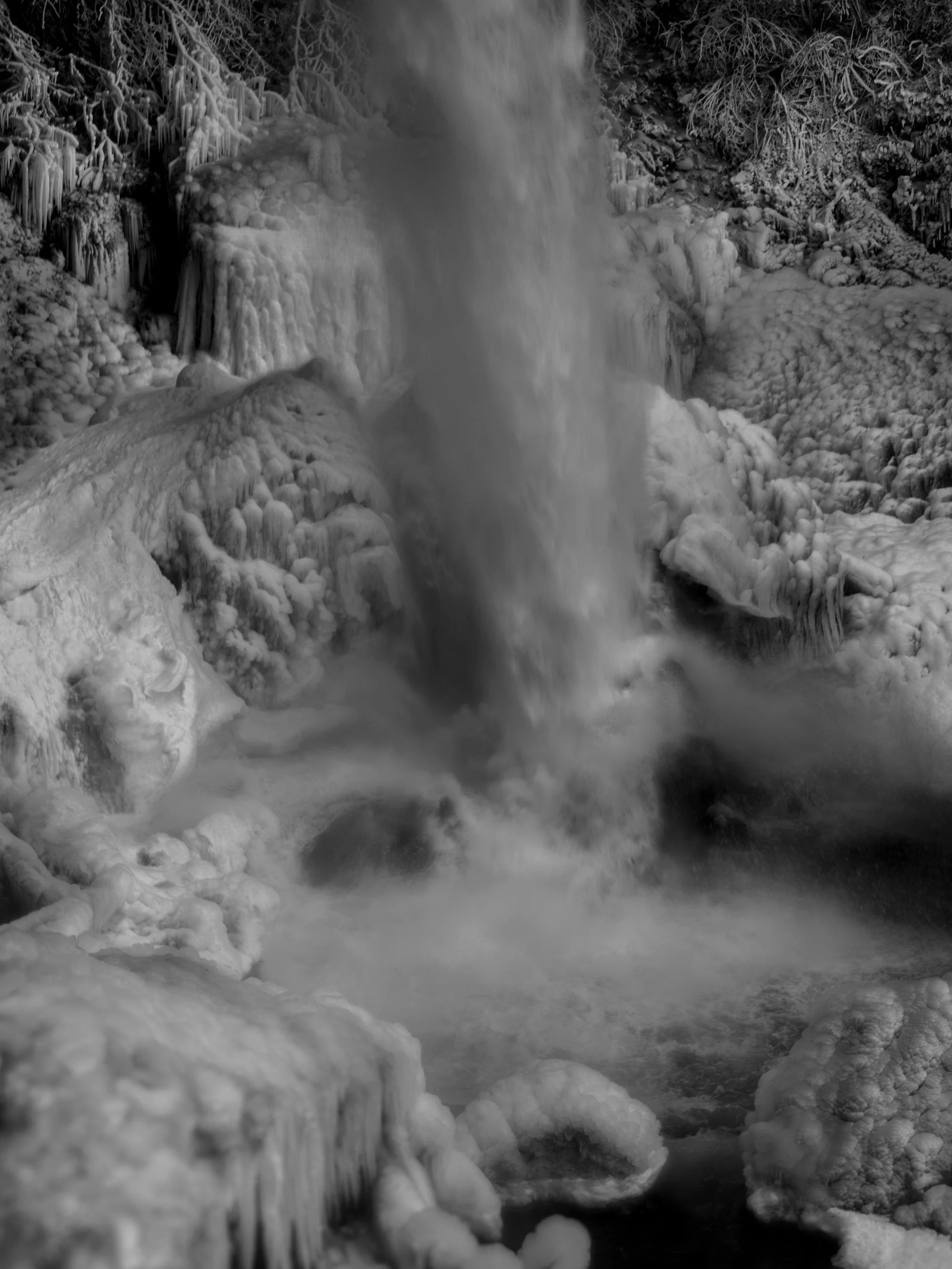 A frozen waterfall with snow and ice covering rocks and trees around it, in a cold winter landscape.