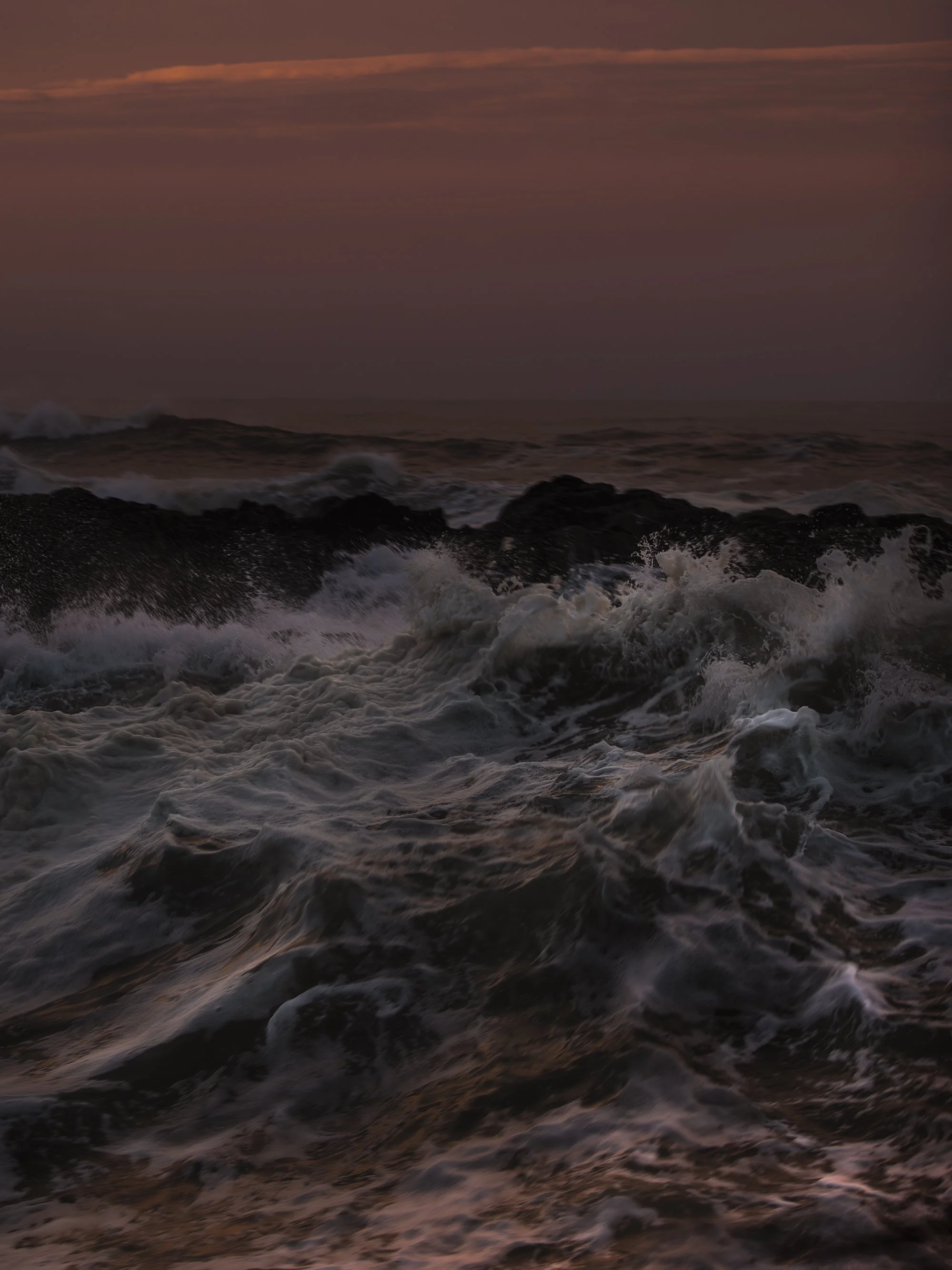Waves crashing against rocks at sunset or sunrise with a dark sky and pinkish clouds overhead.