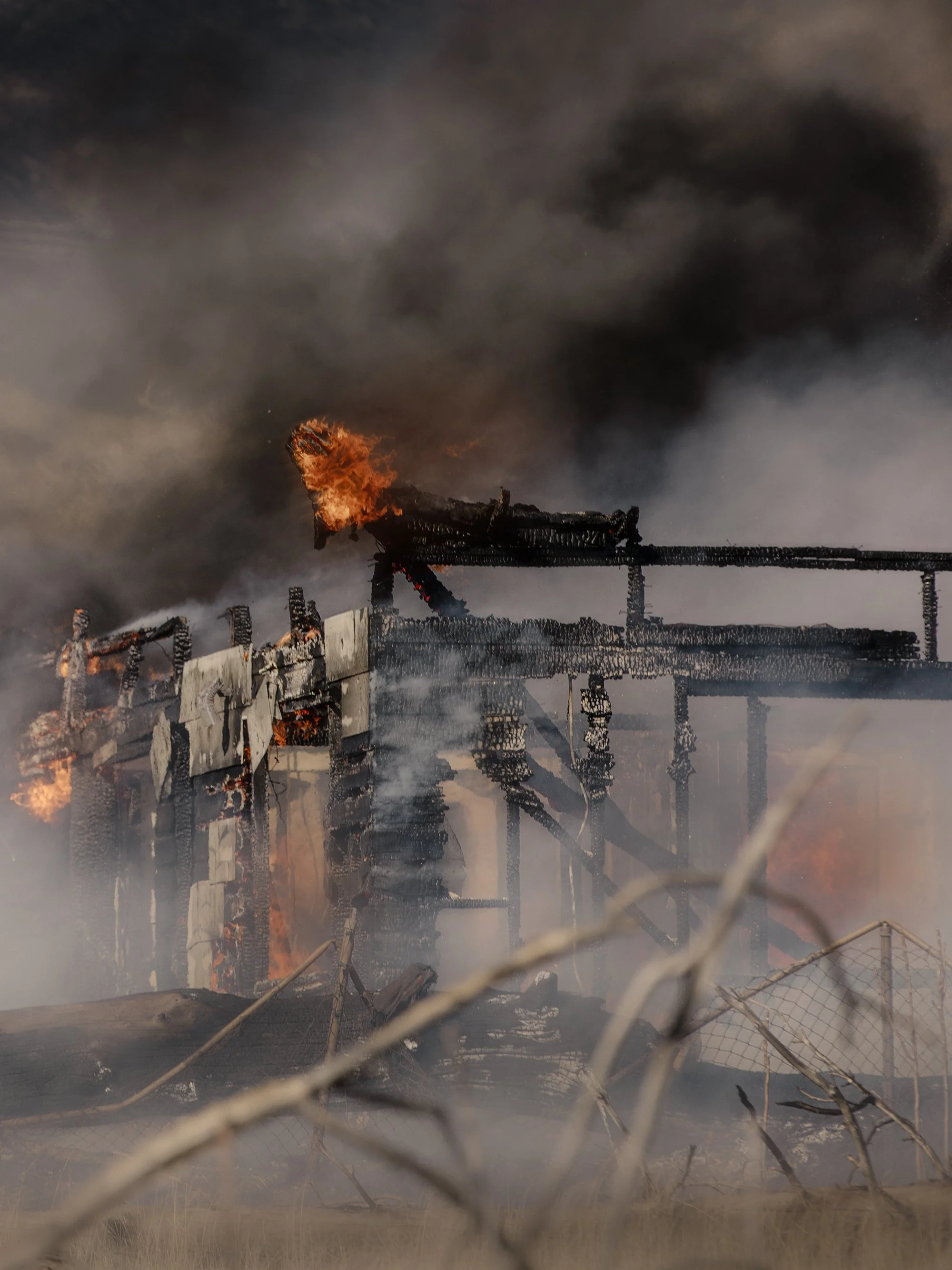 Burning and collapsed house with dark smoke rising into the sky, viewed through a chain-link fence.