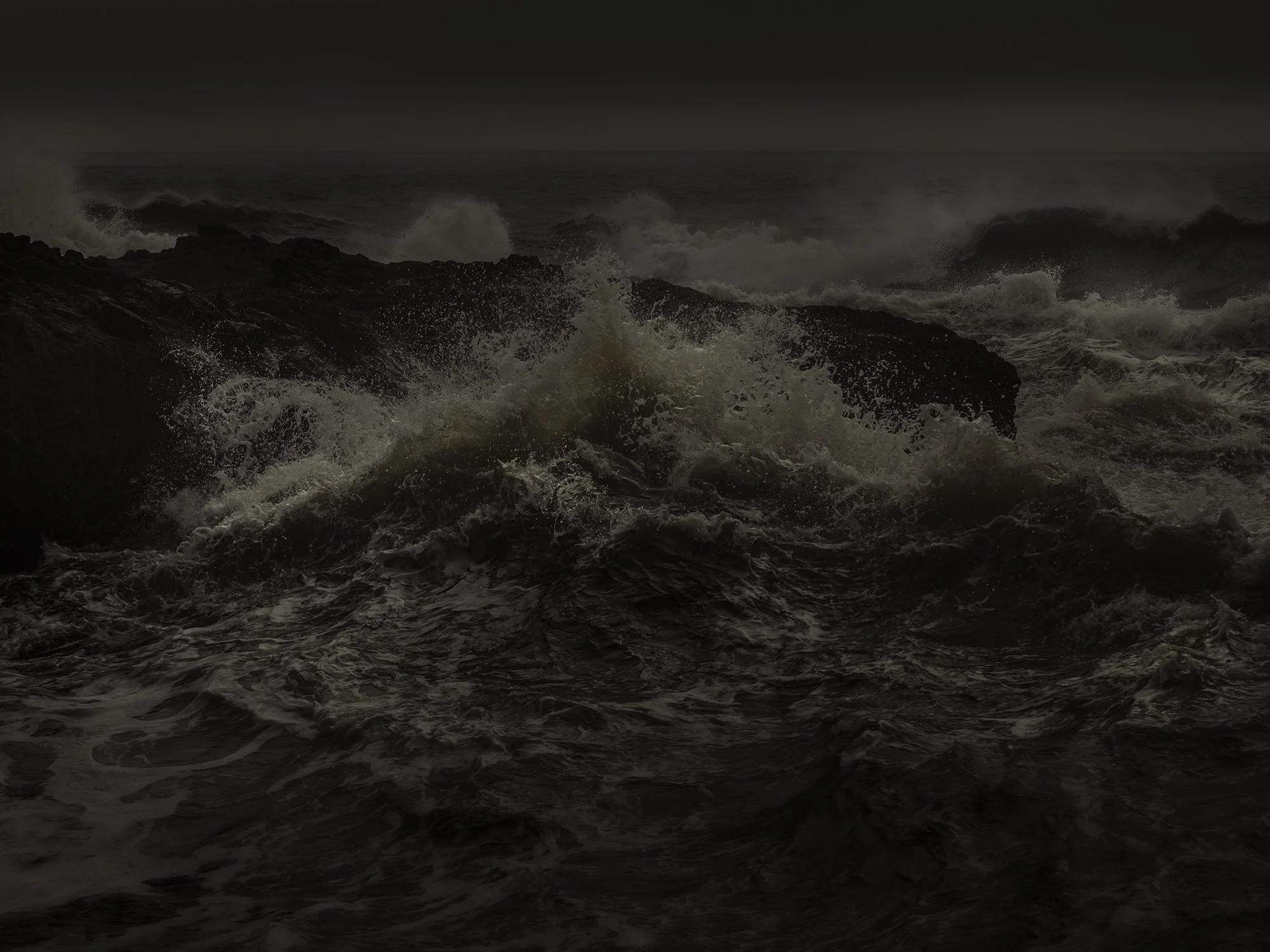 Dark ocean waves crashing against black rocks under a dark, cloudy sky.