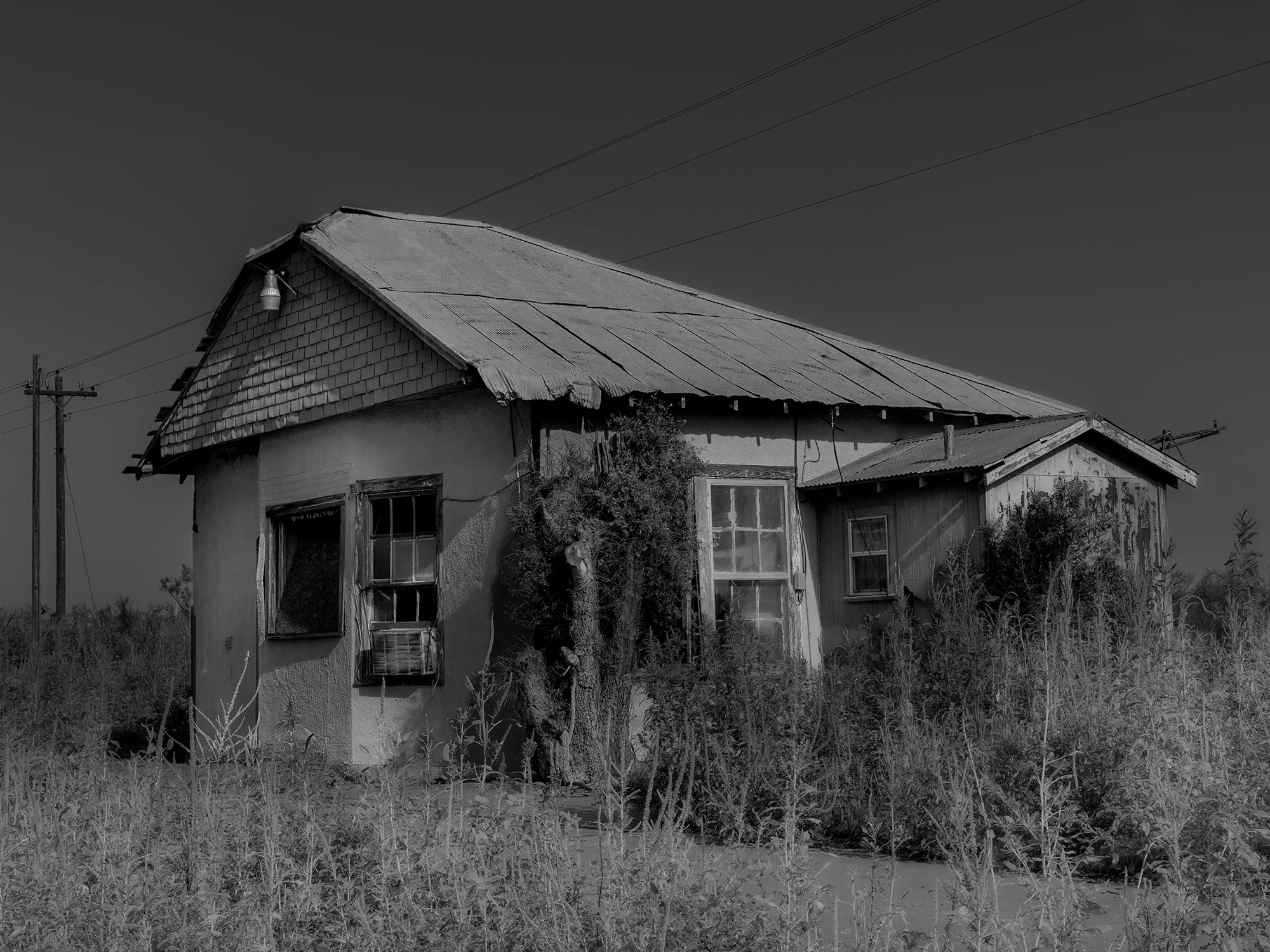 An old, weathered house with a corrugated metal roof and wooden siding, surrounded by overgrown grass and debris, under a clear sky with power lines overhead.