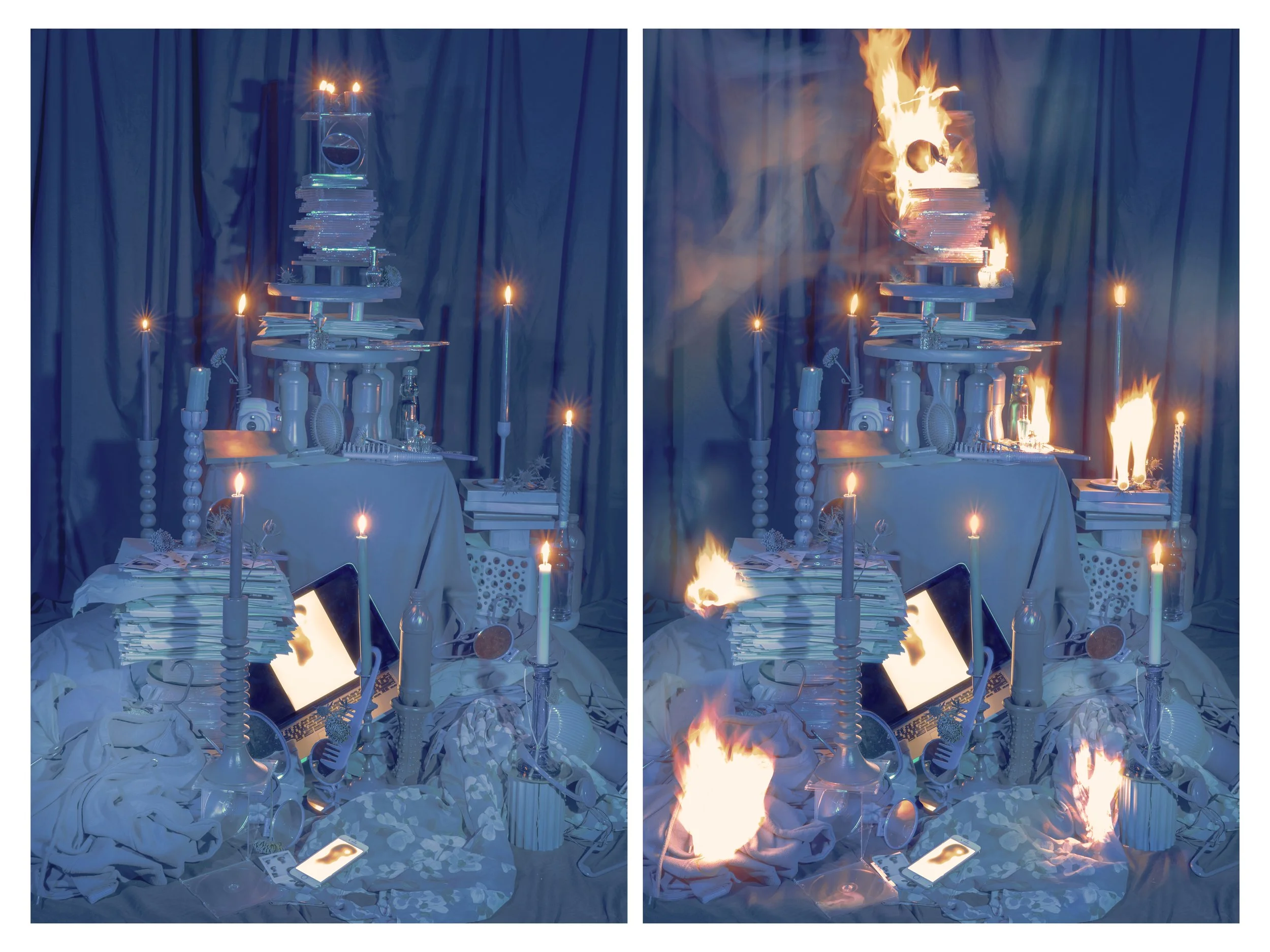An art installation with a multi-tiered table made of stacked plates, books, and bottles, decorated with candles, some of which are on fire, and electronic devices, set against a dark backdrop with blue curtains.