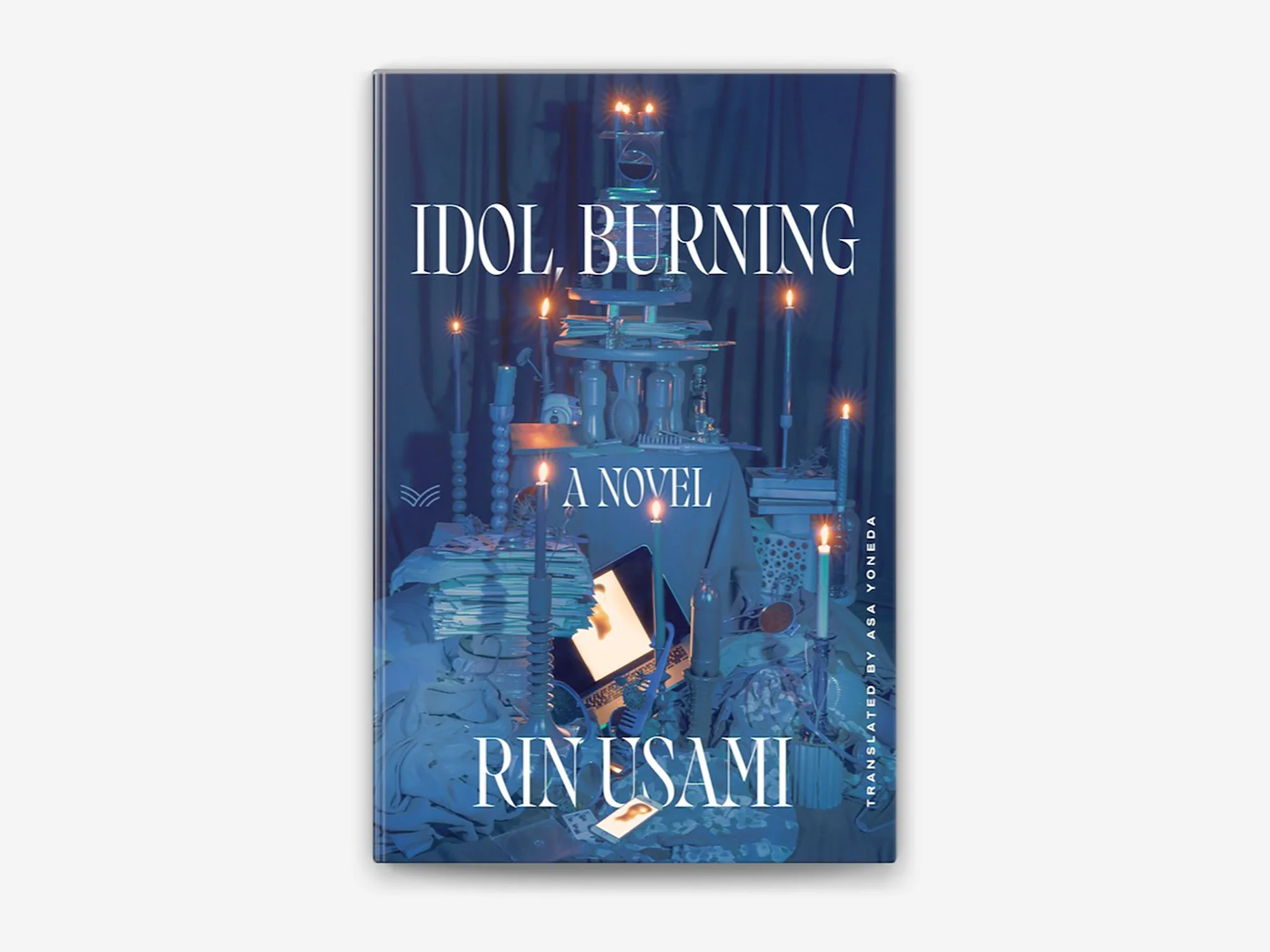 Cover of a novel titled 'Idol, Burning' by Bessy Rinésami, featuring a blue-toned image of a table with candles, books, and various objects, set against dark curtains.