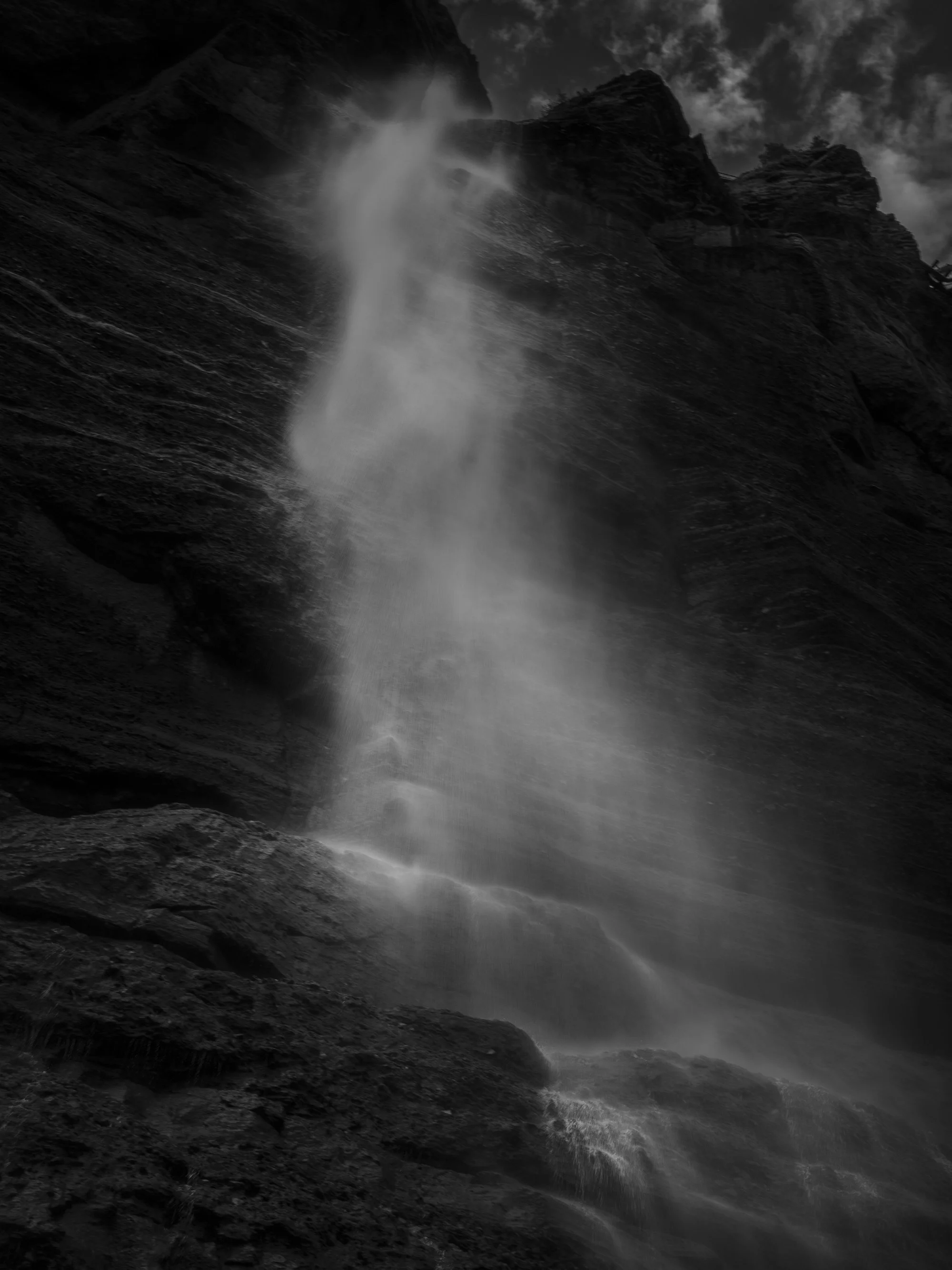 Black and white photo of a waterfall cascading down a steep rocky cliff with clouds in the sky above.