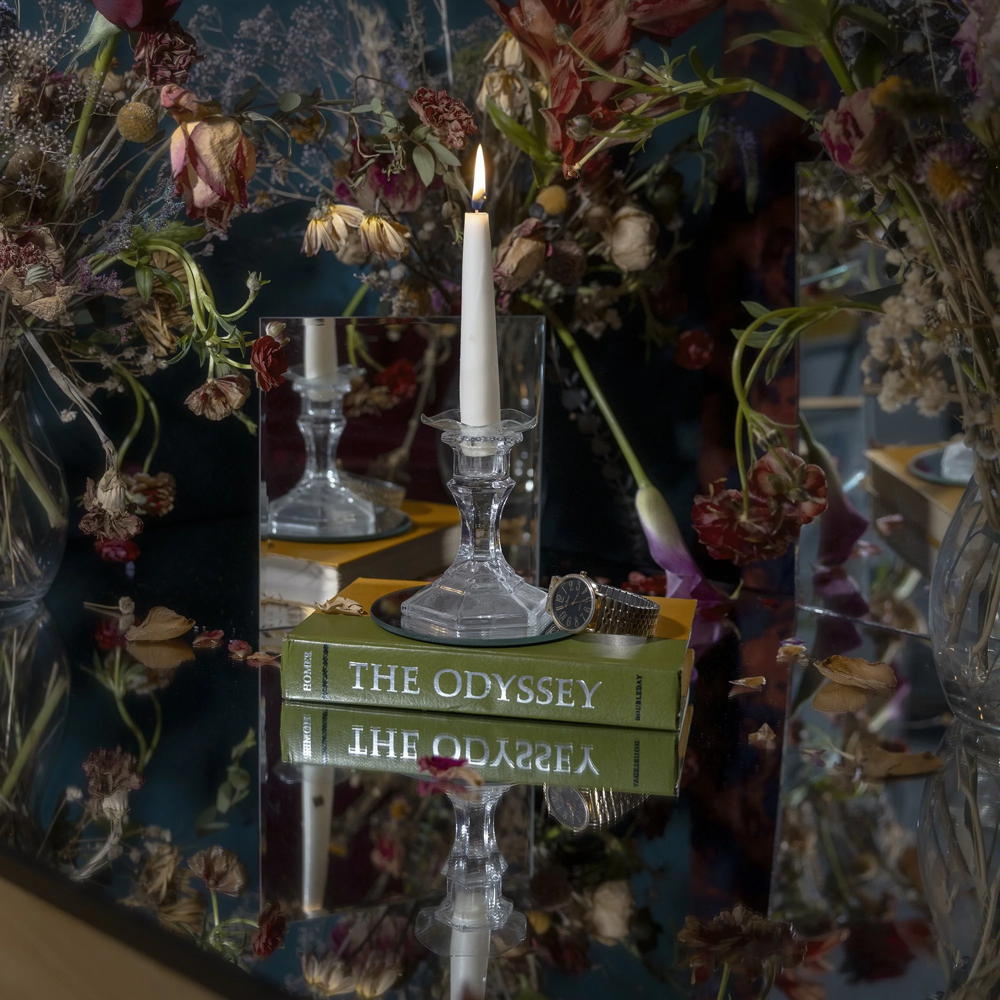 A white candle in a glass holder on top of a stack of green books titled 'The Odyssey' with dried flowers and plants in the background.