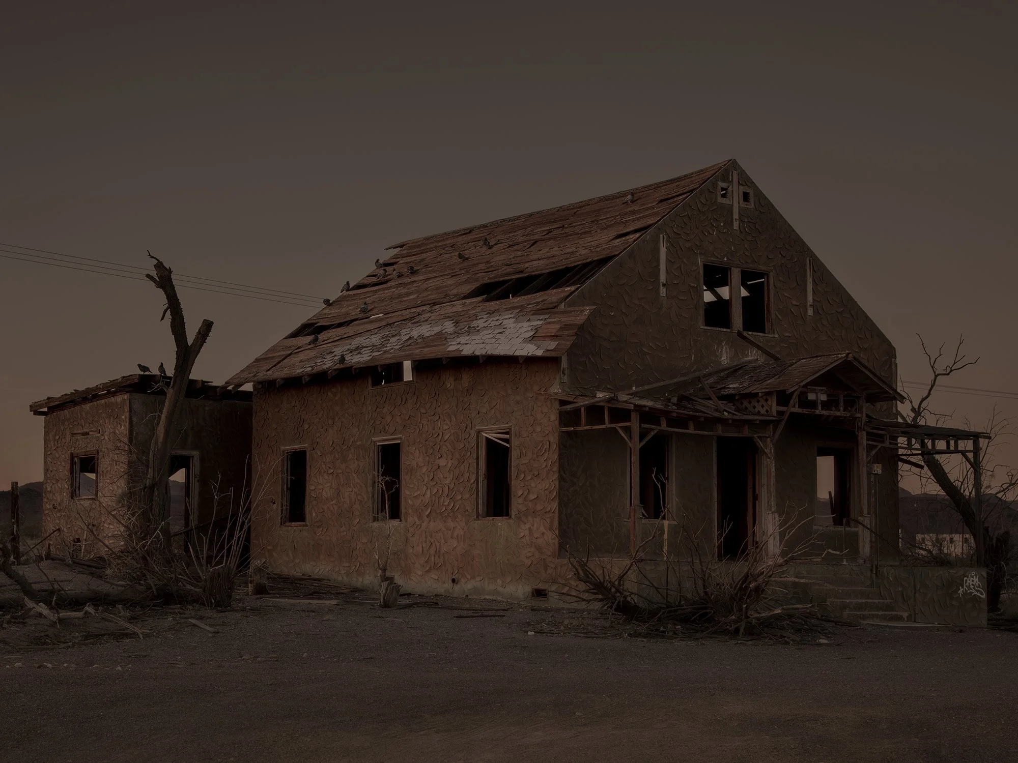 An abandoned, dilapidated house with broken windows and a deteriorated roof, surrounded by dead trees and debris, under a dark, overcast sky.