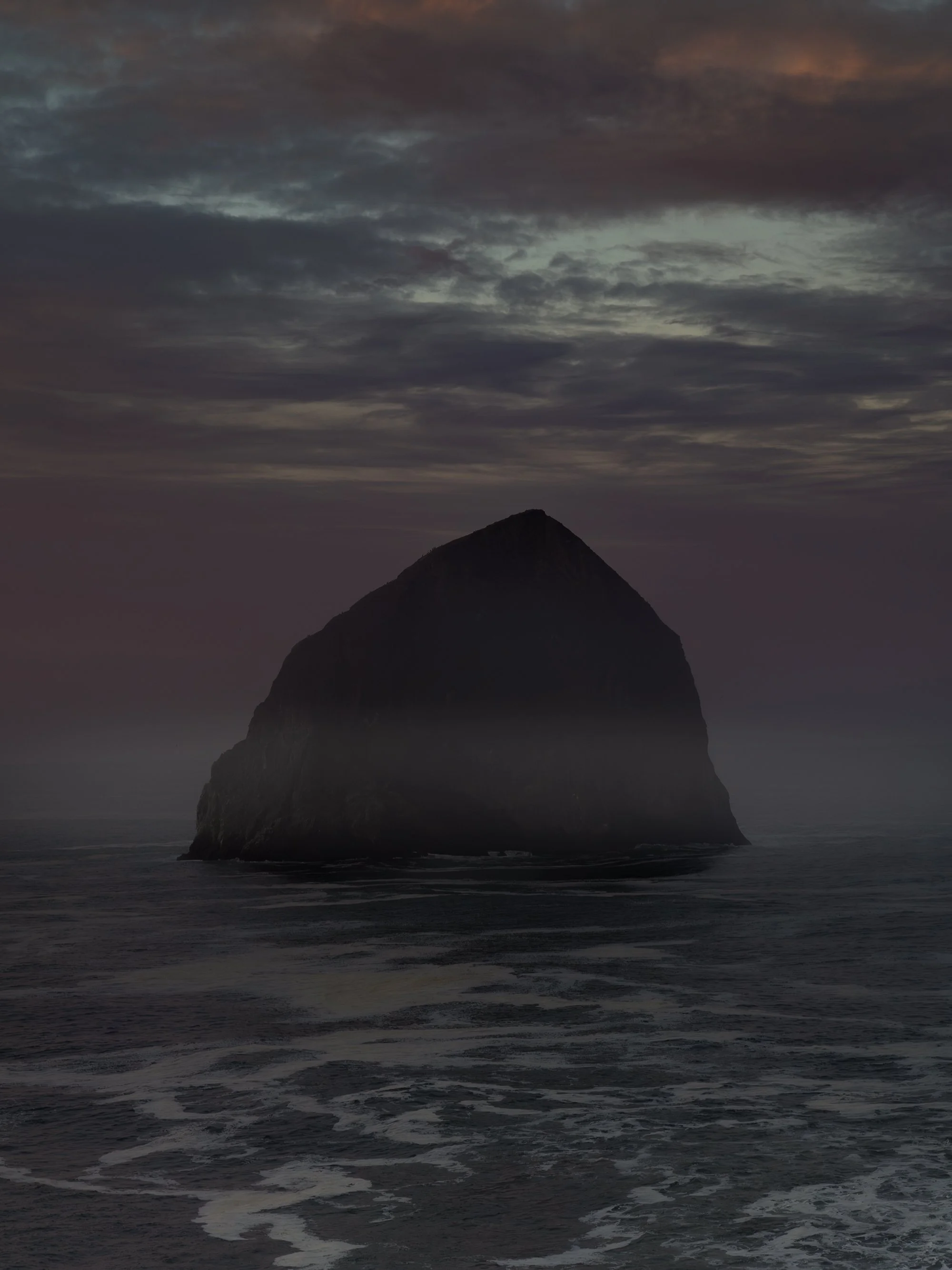 A large rocky sea stack rising from the ocean, surrounded by choppy dark waters, under a cloudy, overcast sky at sunset.